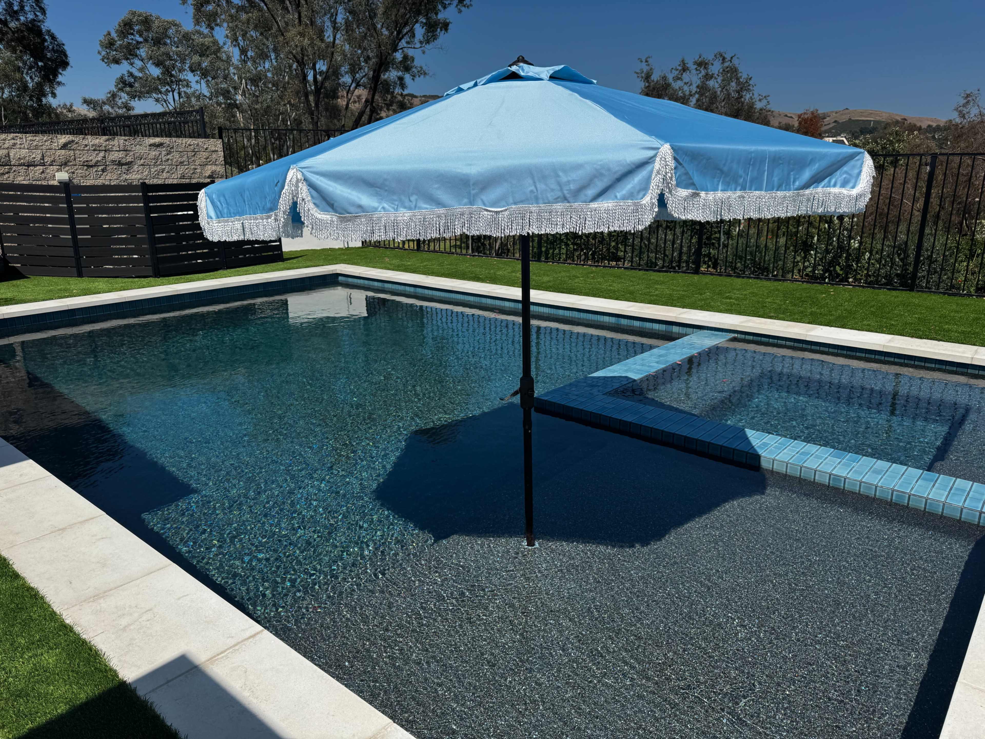 A blue patio umbrella shades a section of a swimming pool surrounded by green grass and a black fence.