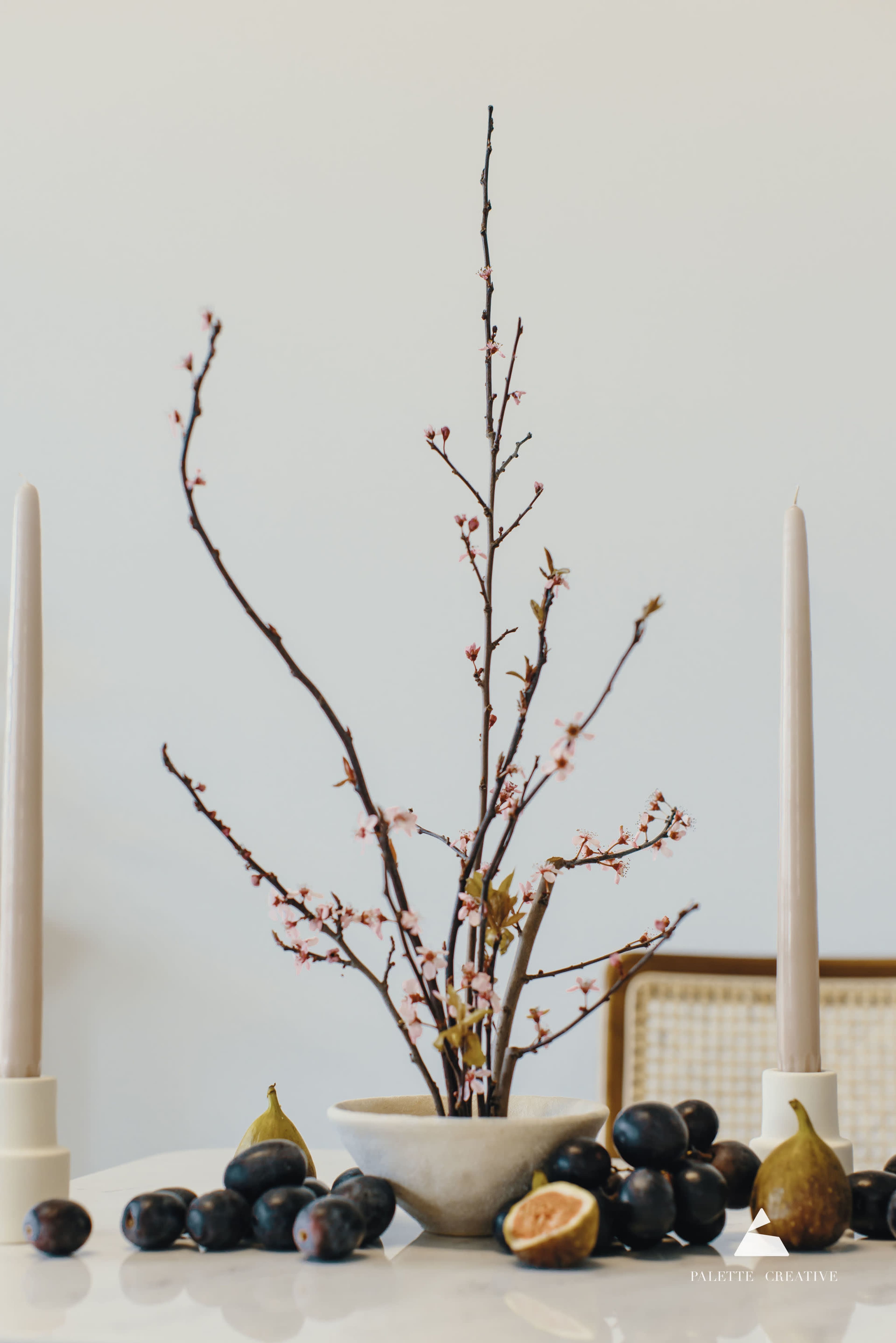 A branch with pink blossoms is showcased in a vase on a table, accompanied by two candles and assorted fruits, including figs and plums.