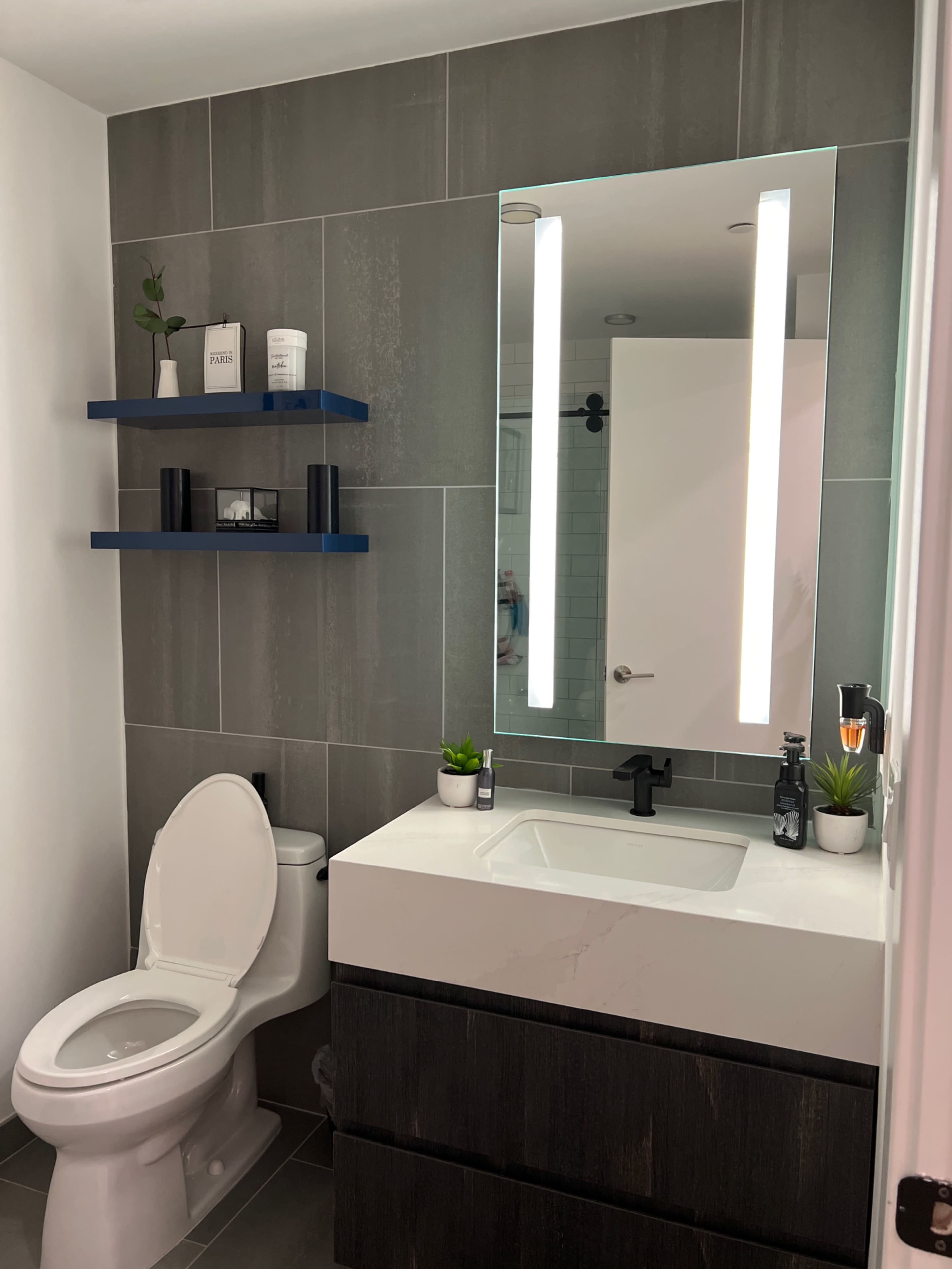 A modern bathroom features a wall-mounted mirror with LED lights above a rectangular sink and dark cabinetry, alongside a toilet and open shelves displaying decorative items.