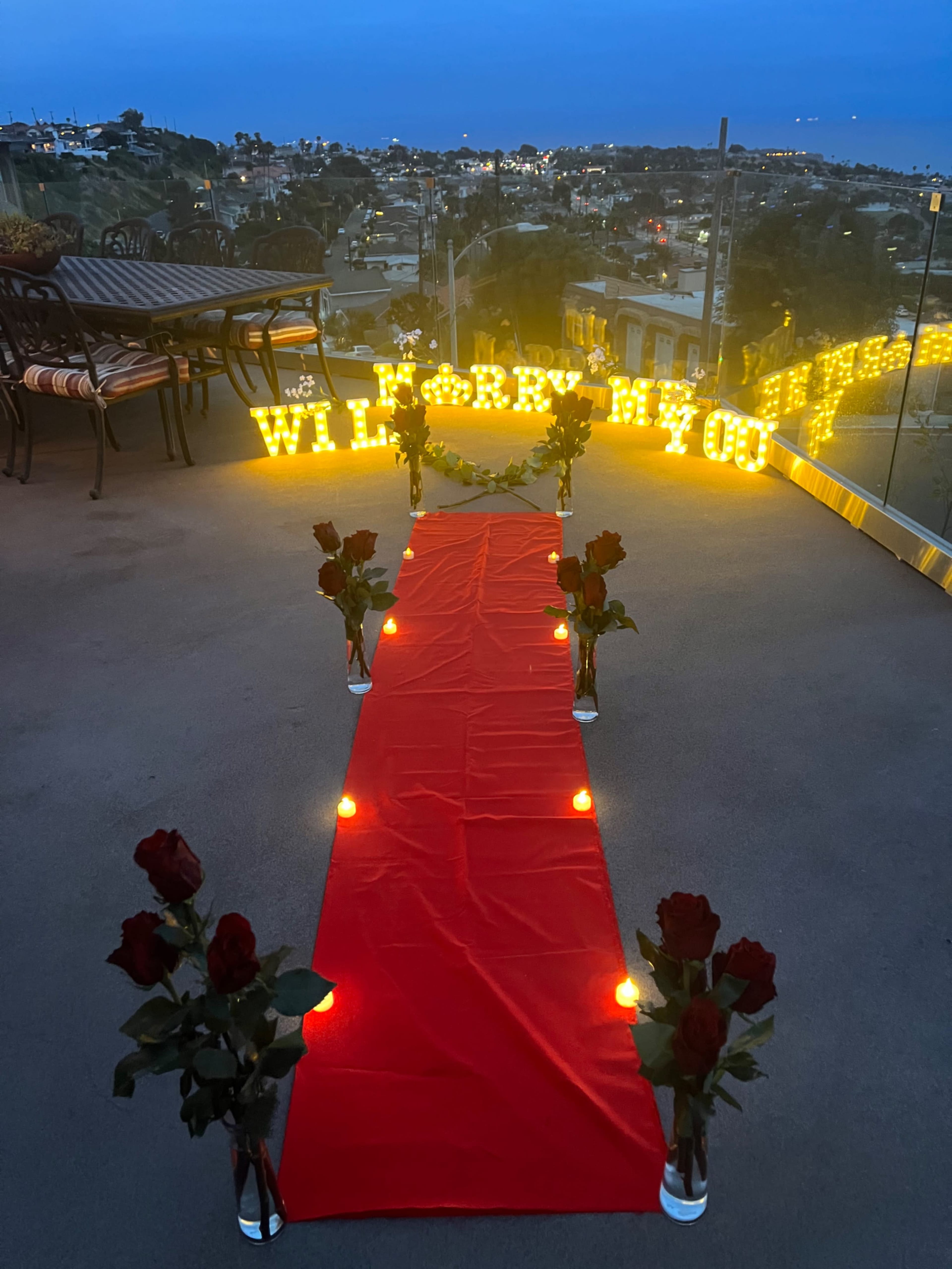 A red carpet lined with candles and roses leads to illuminated letters that read "WILL YOU MARRY ME" on a balcony overlooking a city at dusk.