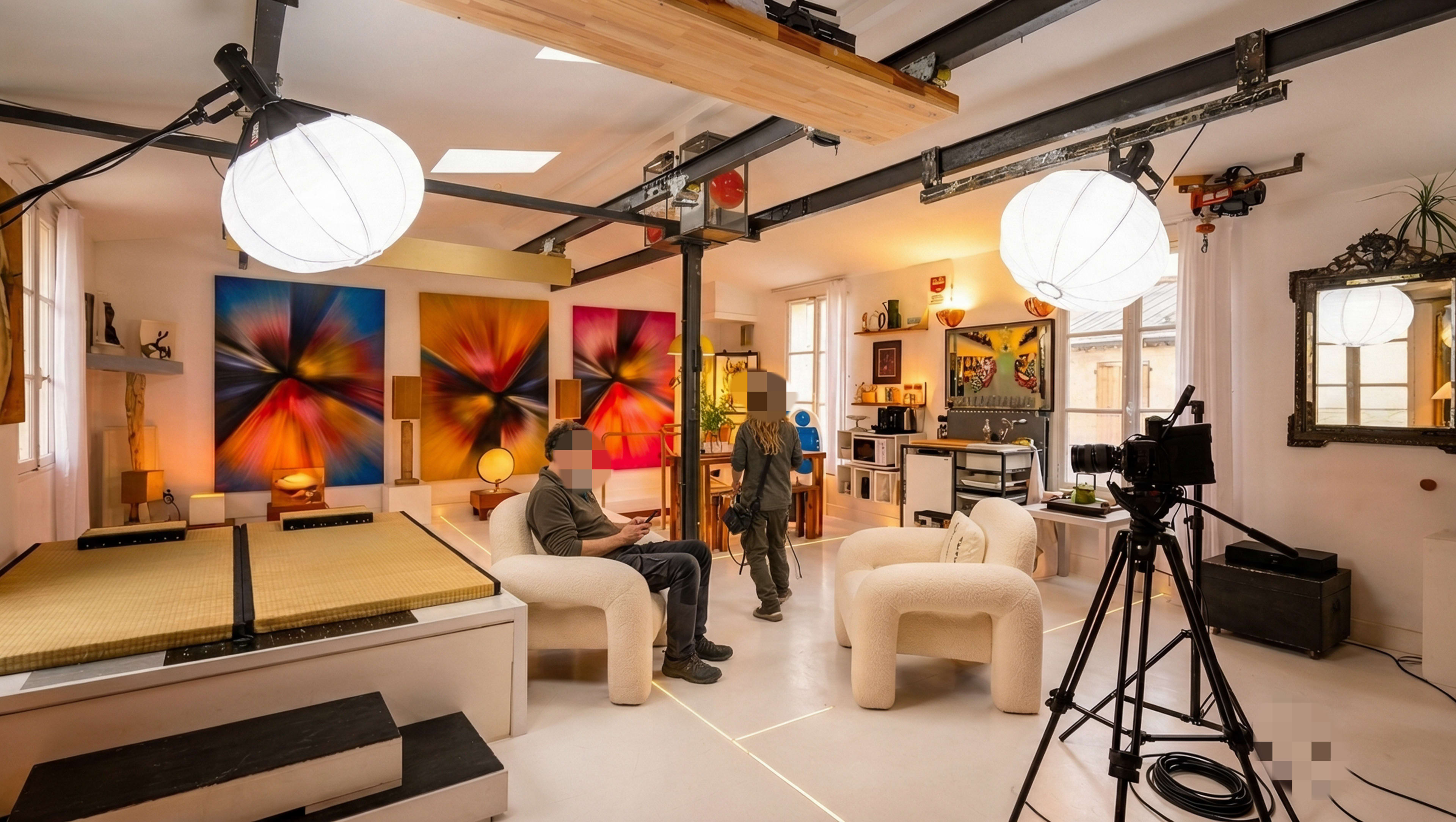 The image shows a modern, well-lit interior space featuring two chairs, a camera setup, vibrant artwork on the walls, and various decor items.