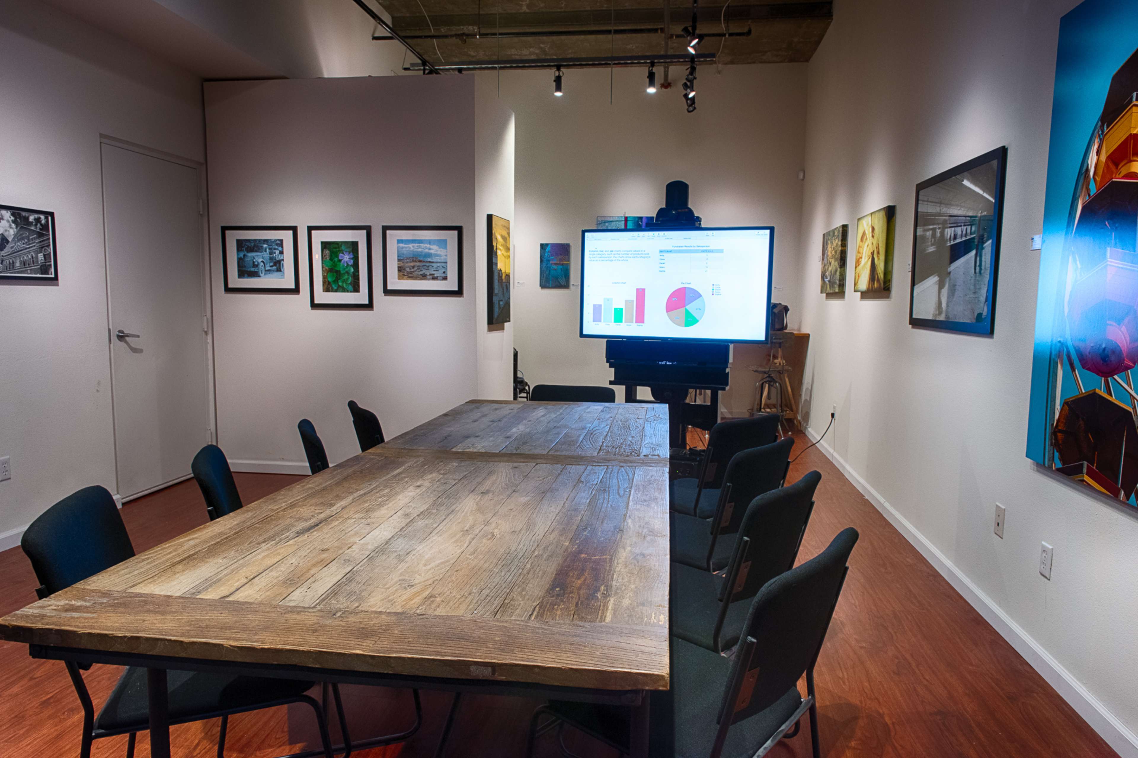 A large wooden table surrounded by black chairs is set in an art-filled room with a presentation screen displaying graphs and charts.