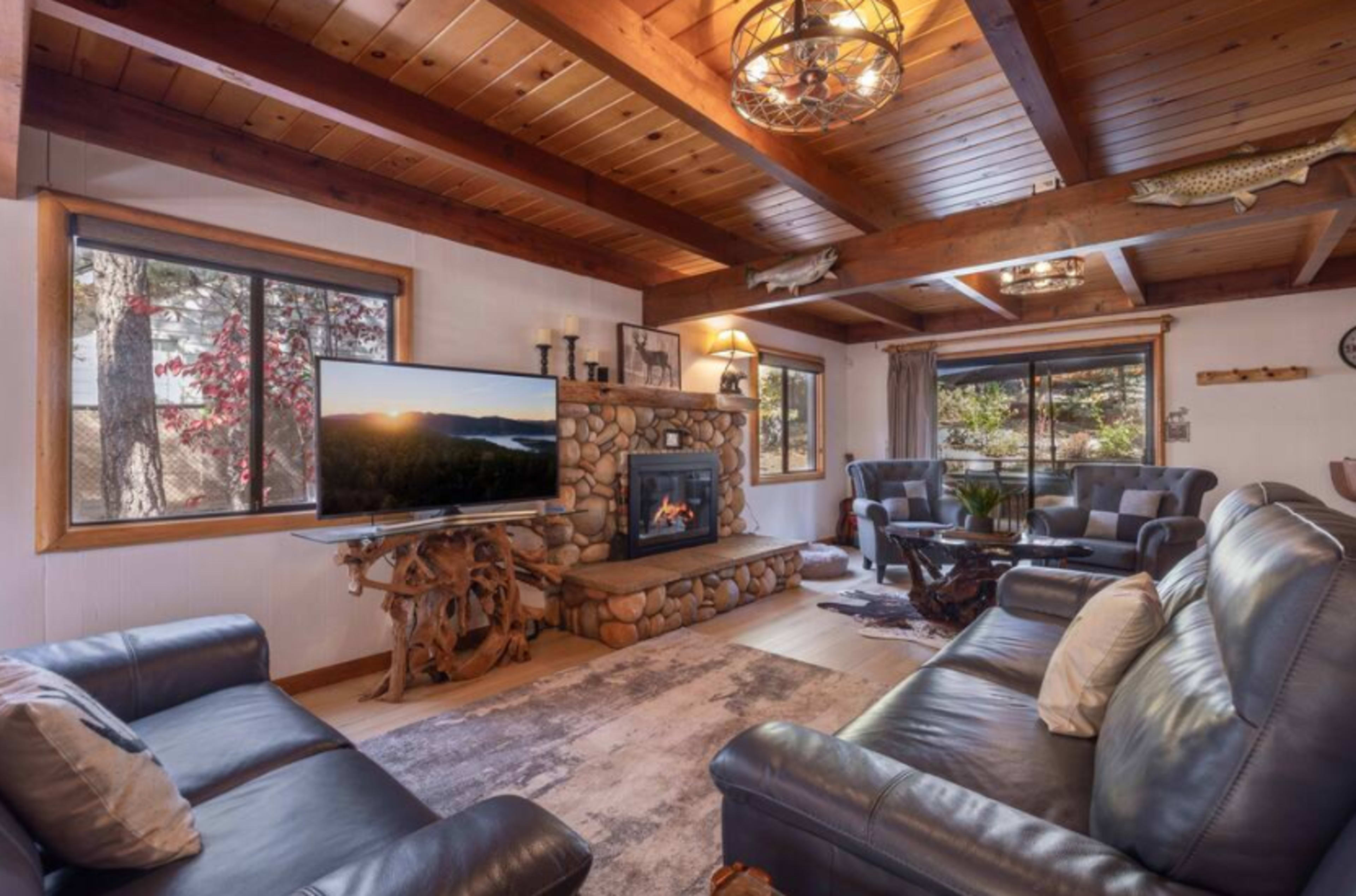 The living room features a stone fireplace, wooden beams, and a television opposite two sofas, with large windows allowing natural light.