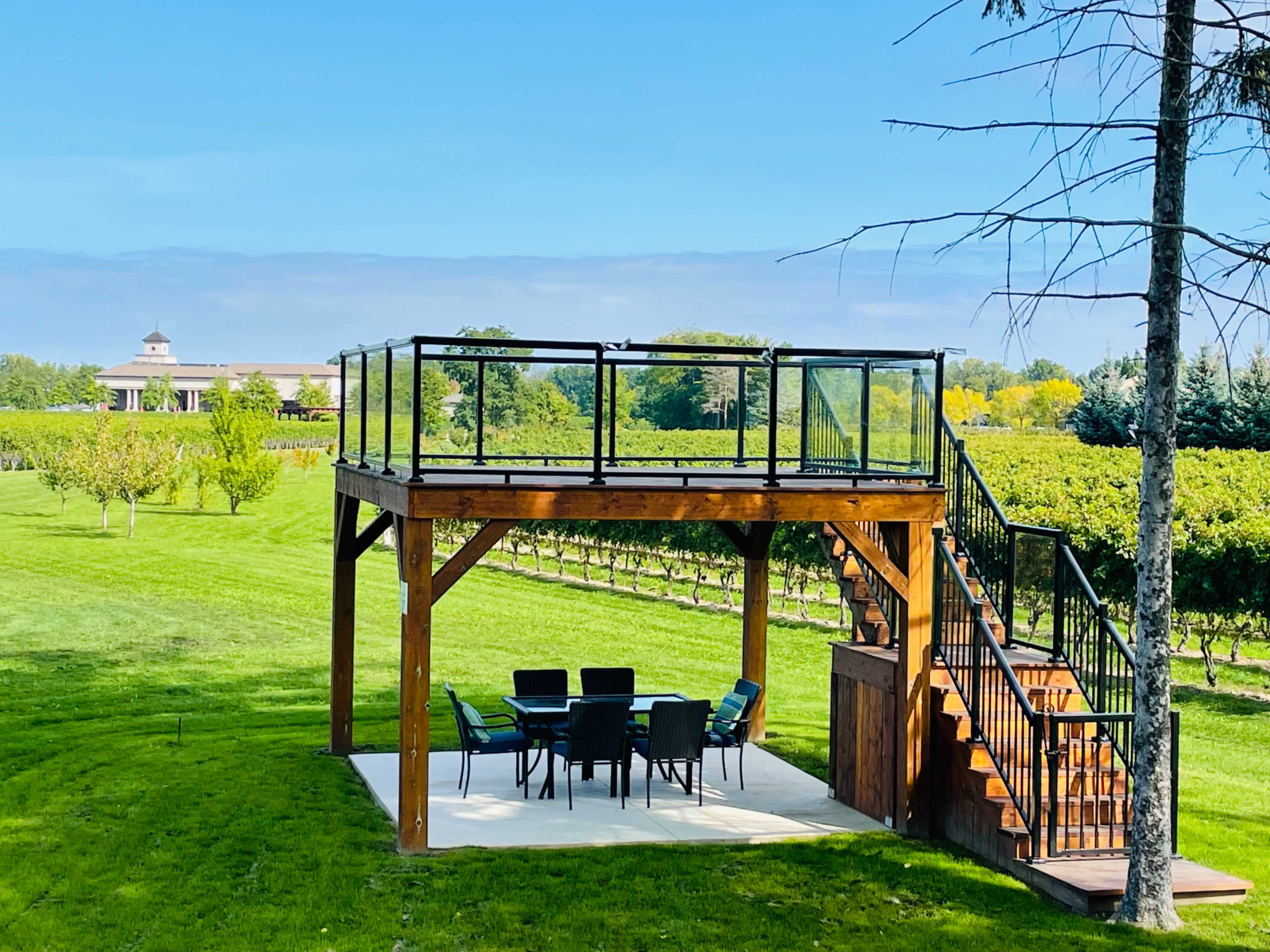 A wooden elevated deck with a glass railing overlooks a green vineyard and is connected by stairs to a patio area with a dining set.
