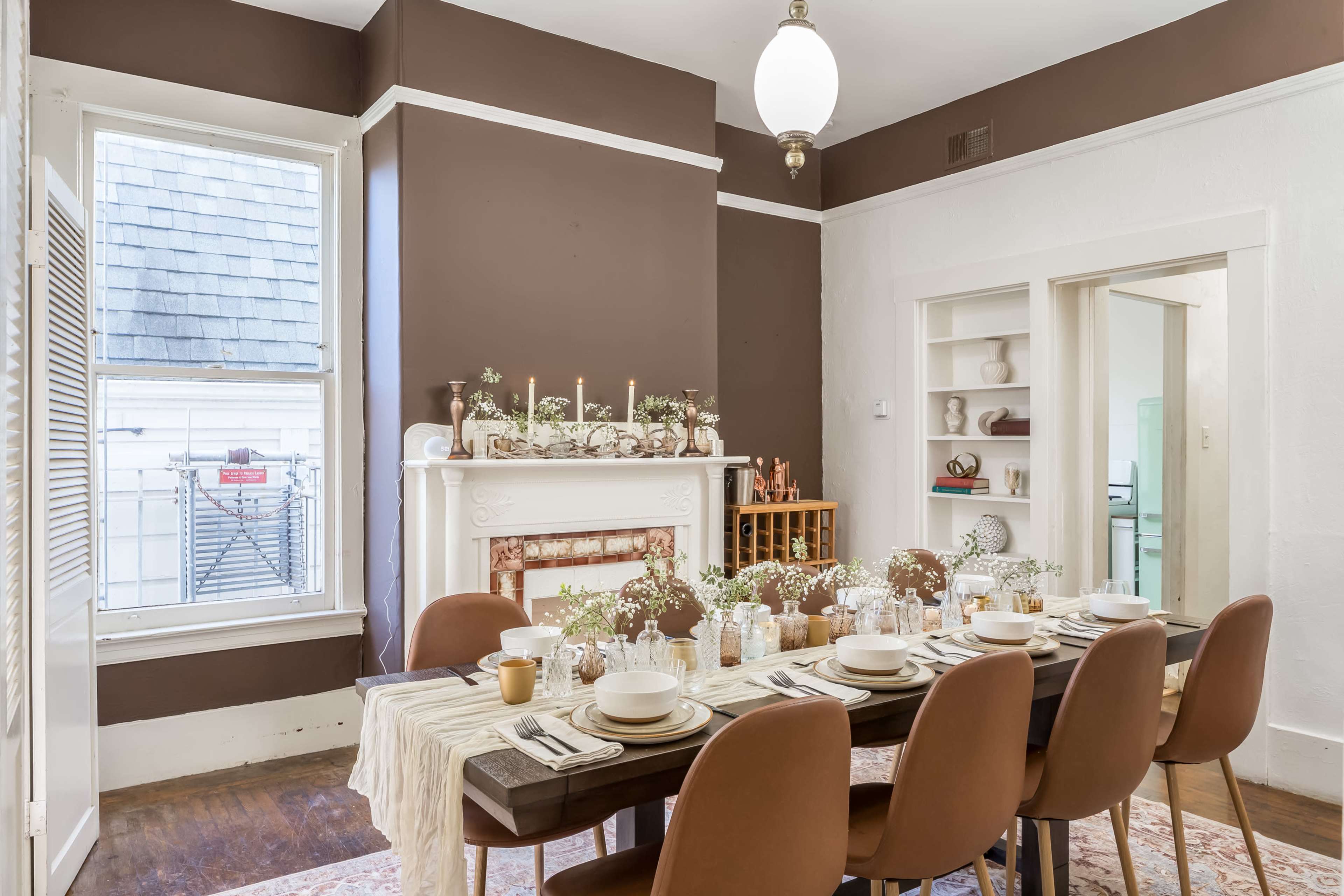 A dining room features a long table set for a meal, with a decorative centerpiece and candles, against a backdrop of brown walls and a white fireplace.