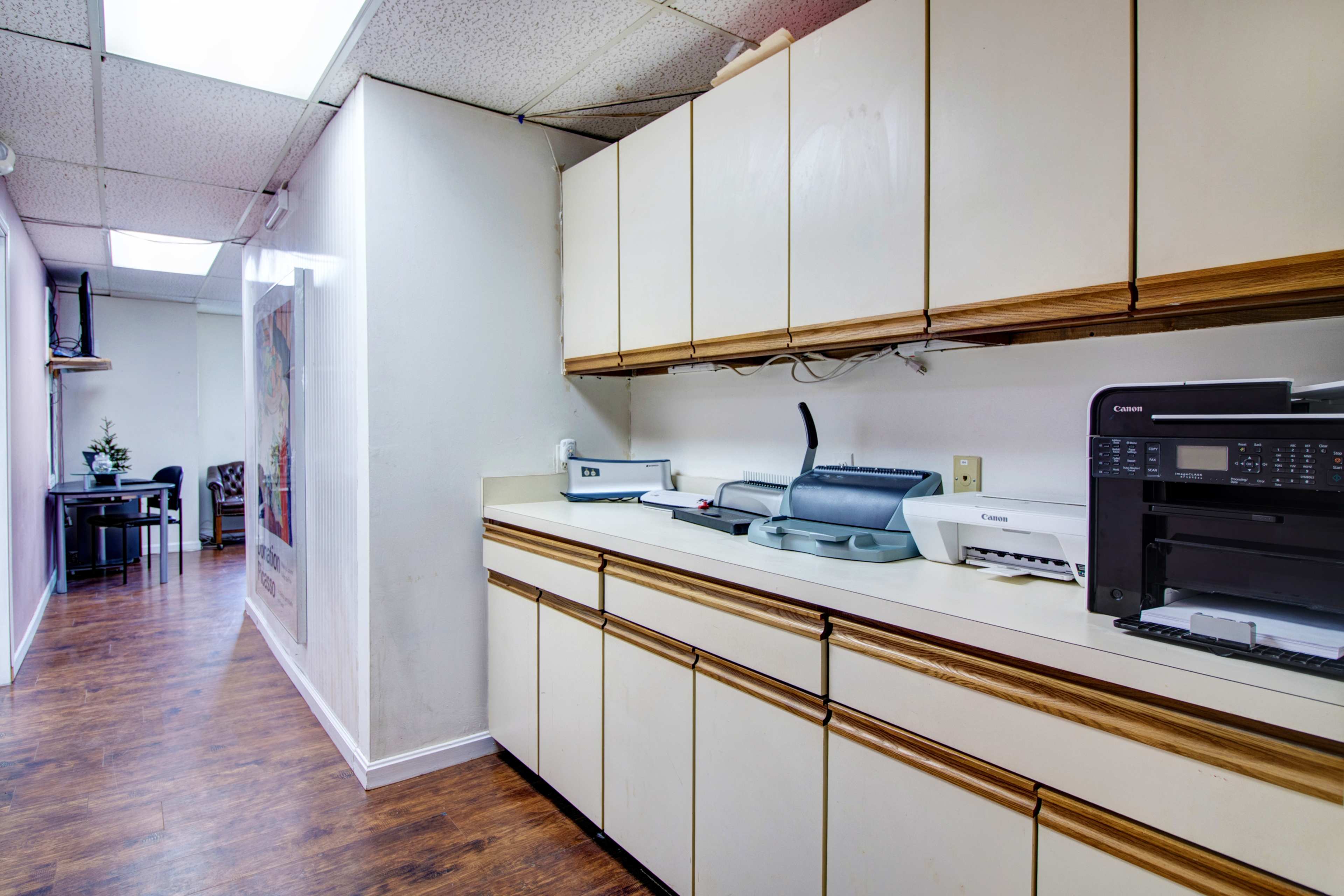 A narrow hallway leads to a kitchen area with beige cabinets, a countertop, and various office equipment, including printers and a scanner.