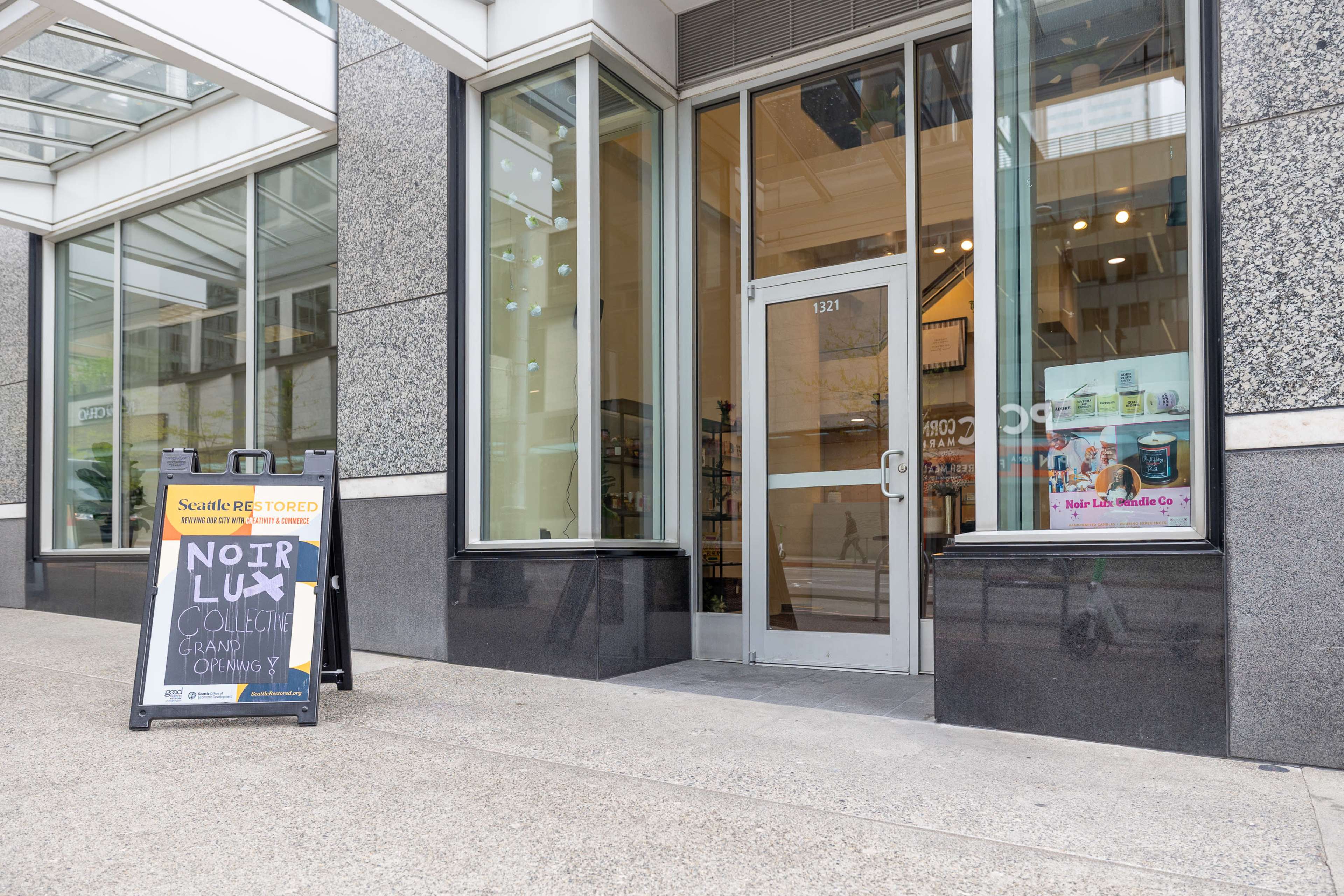 The entrance to a building features a clear glass door, with a sign advertising a "Noir Lux Collective Grand Opening" displayed on an A-frame sign outside.