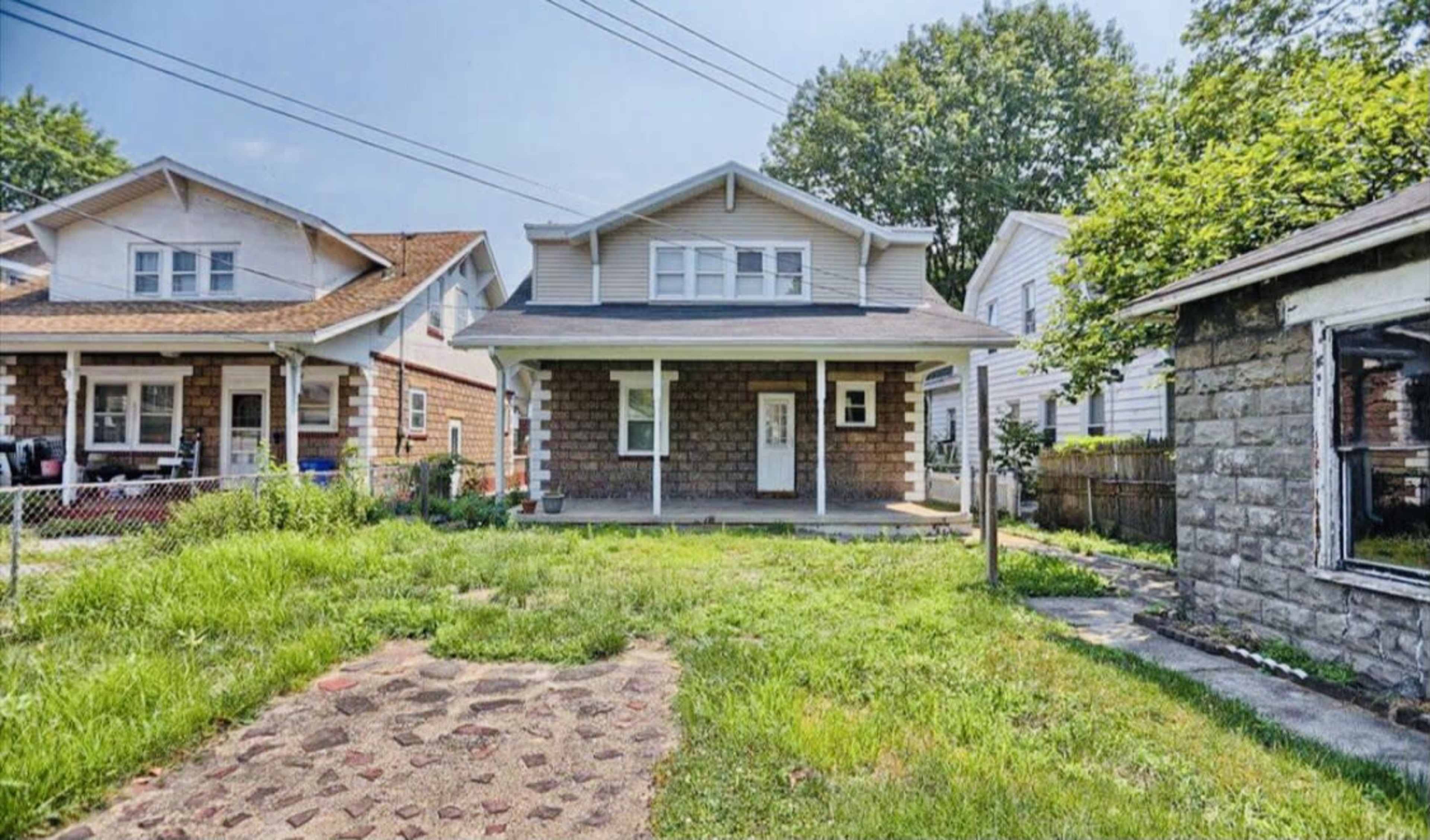 A two-story house with a covered porch, surrounded by overgrown grass and located next to similar residential buildings.