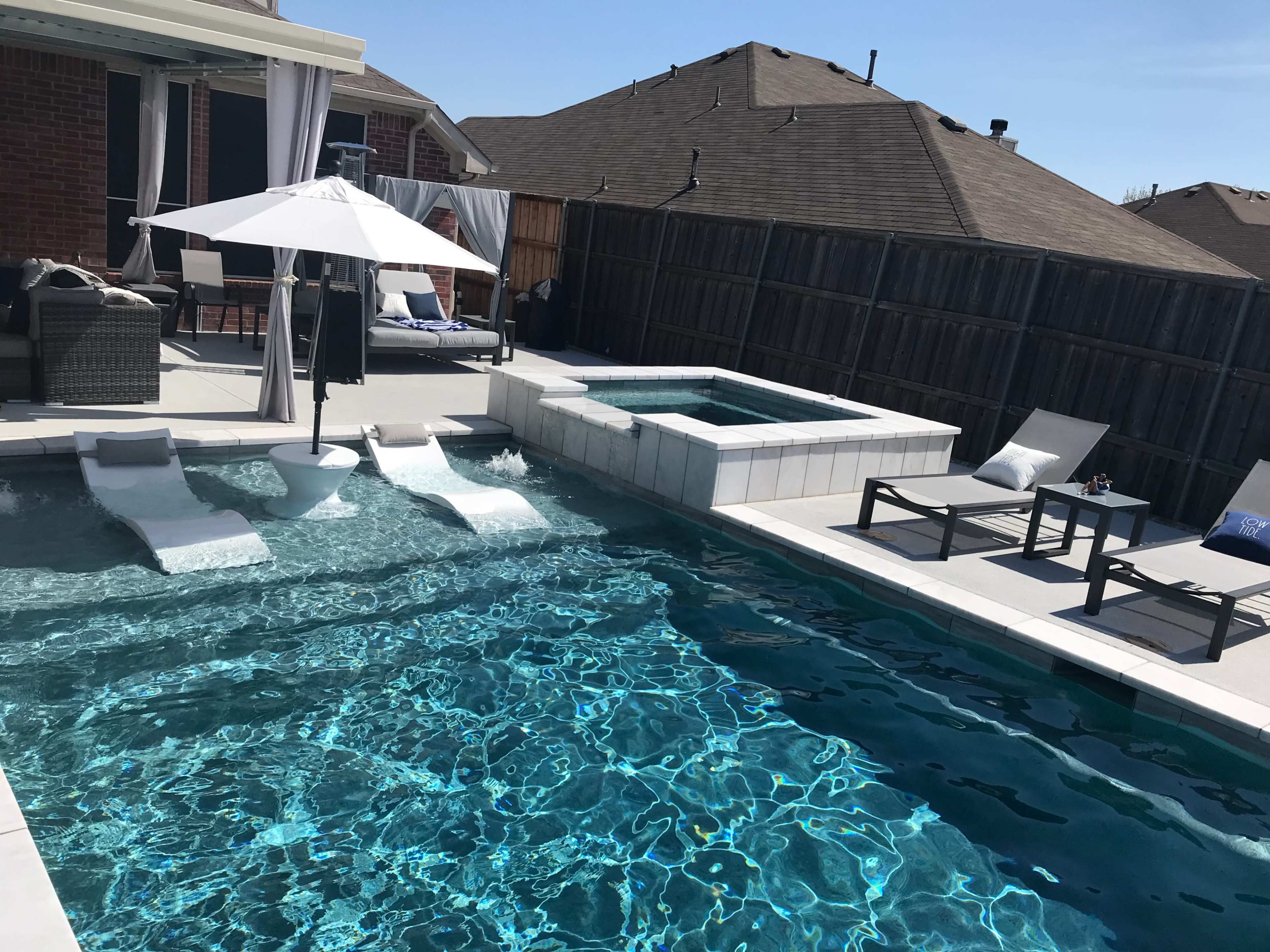 The image shows a backyard pool area with lounge chairs, a hot tub, and a patio umbrella surrounded by a wooden fence.