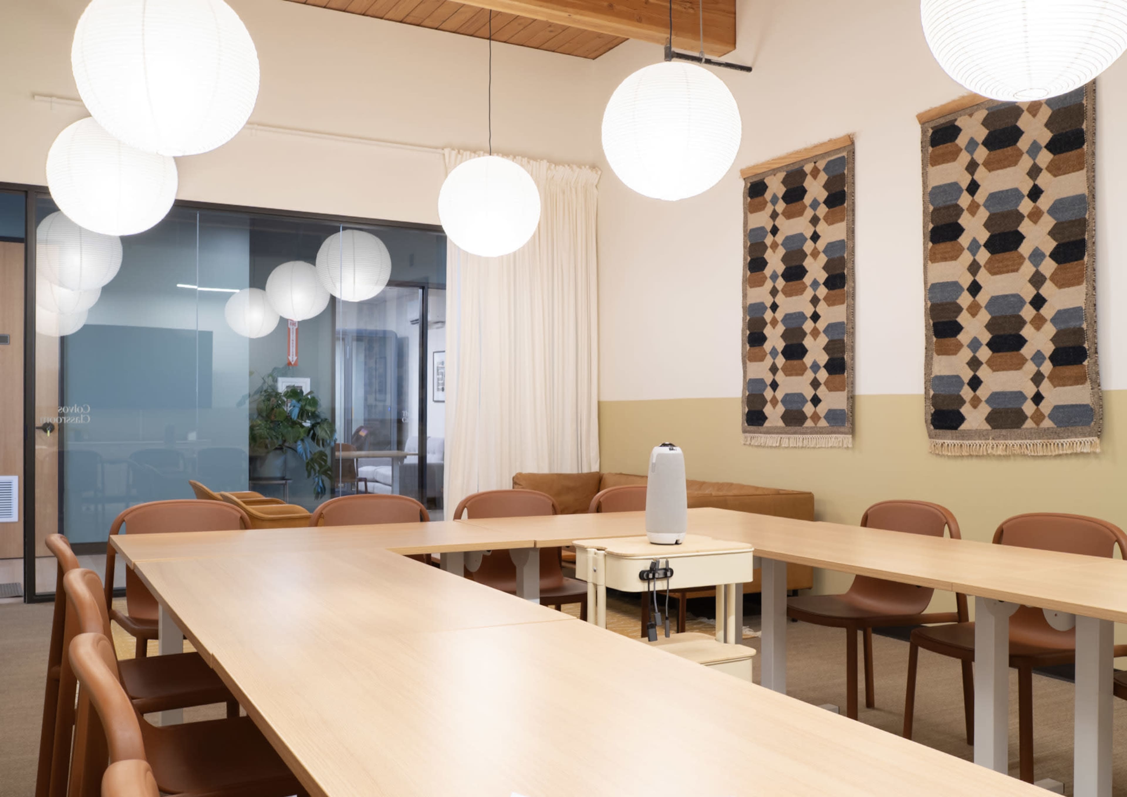 The image shows a modern meeting room with a long table, brown chairs, and decorative wall hangings, illuminated by round pendant lights.