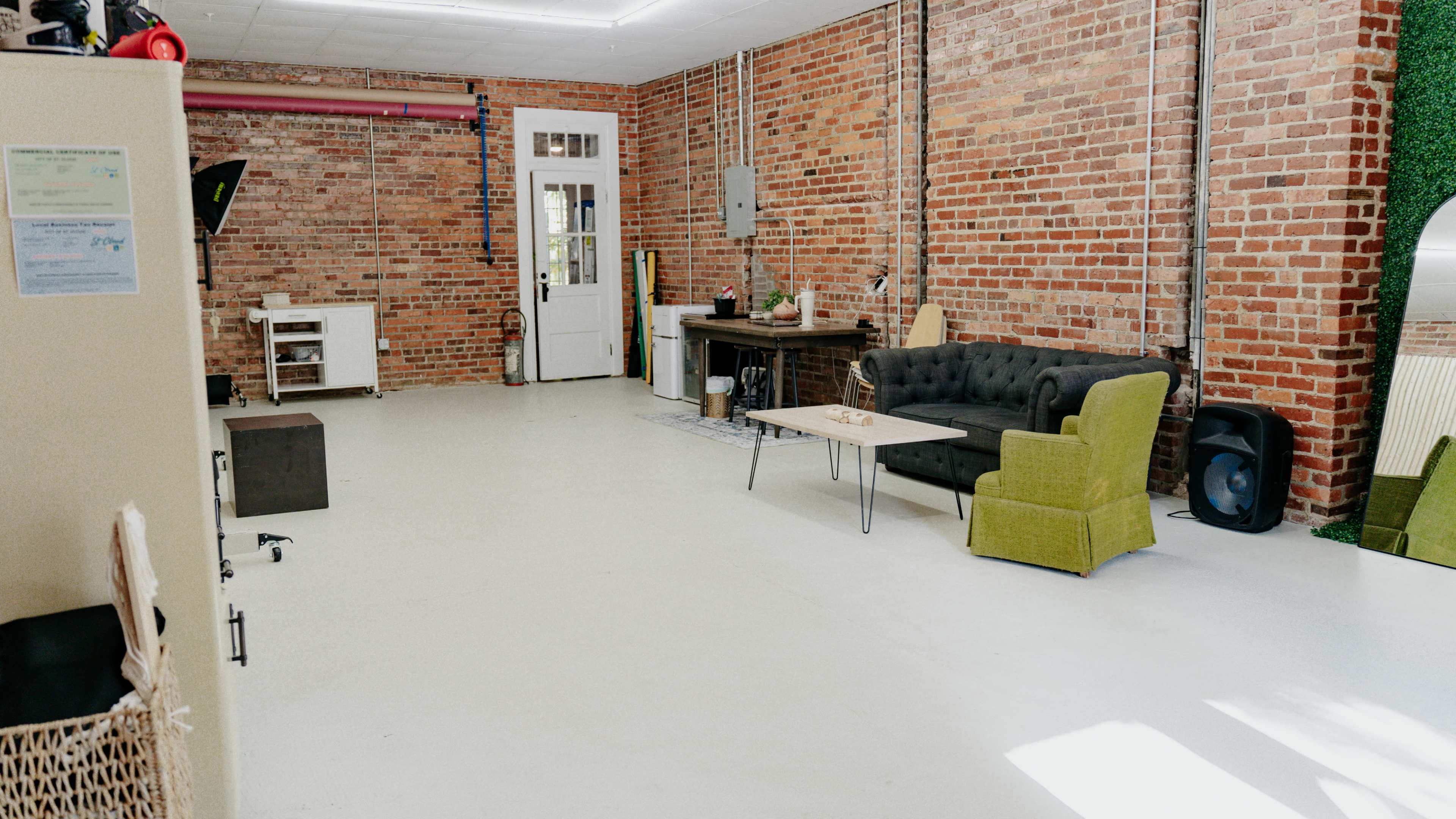 The image shows a spacious room with exposed brick walls, a green armchair, a black couch, a small coffee table, and a doorway leading to another area.