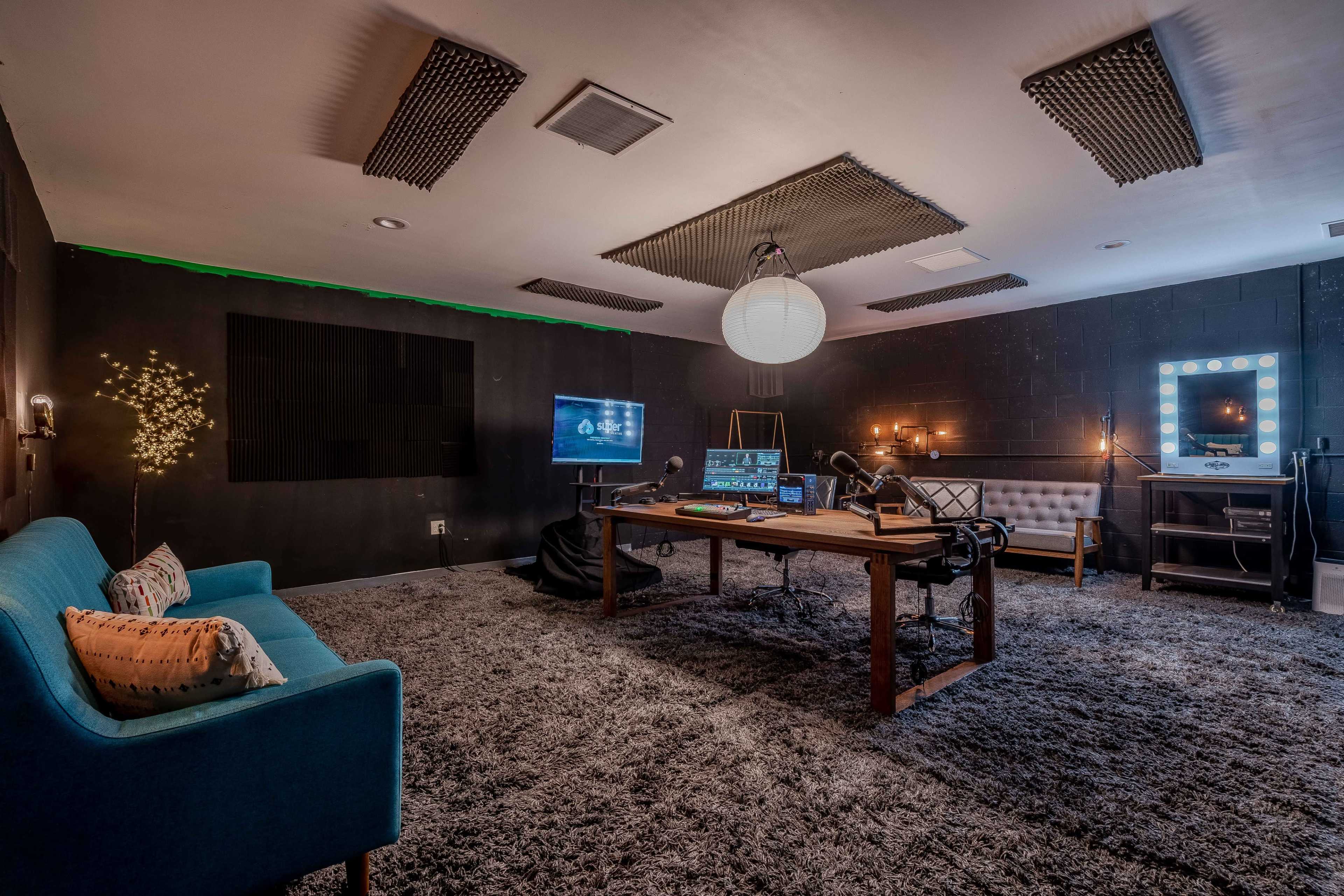 A soundproof studio features a large wooden table, a sofa, and a large mirror with lights, all set against dark walls and a plush carpet.