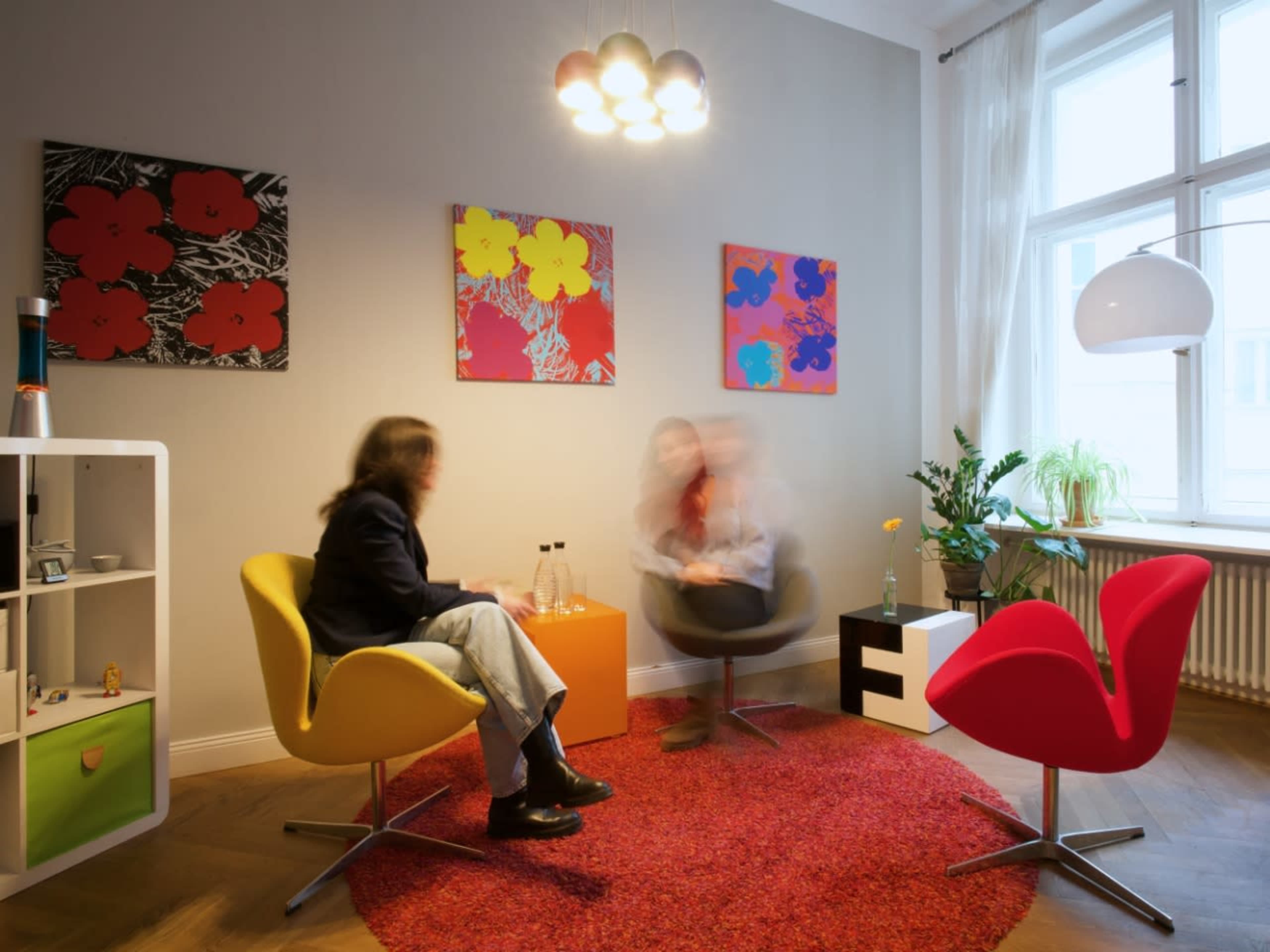 Two people are seated in colorful chairs in a modern room with abstract artwork on the walls and a round red rug on the floor.