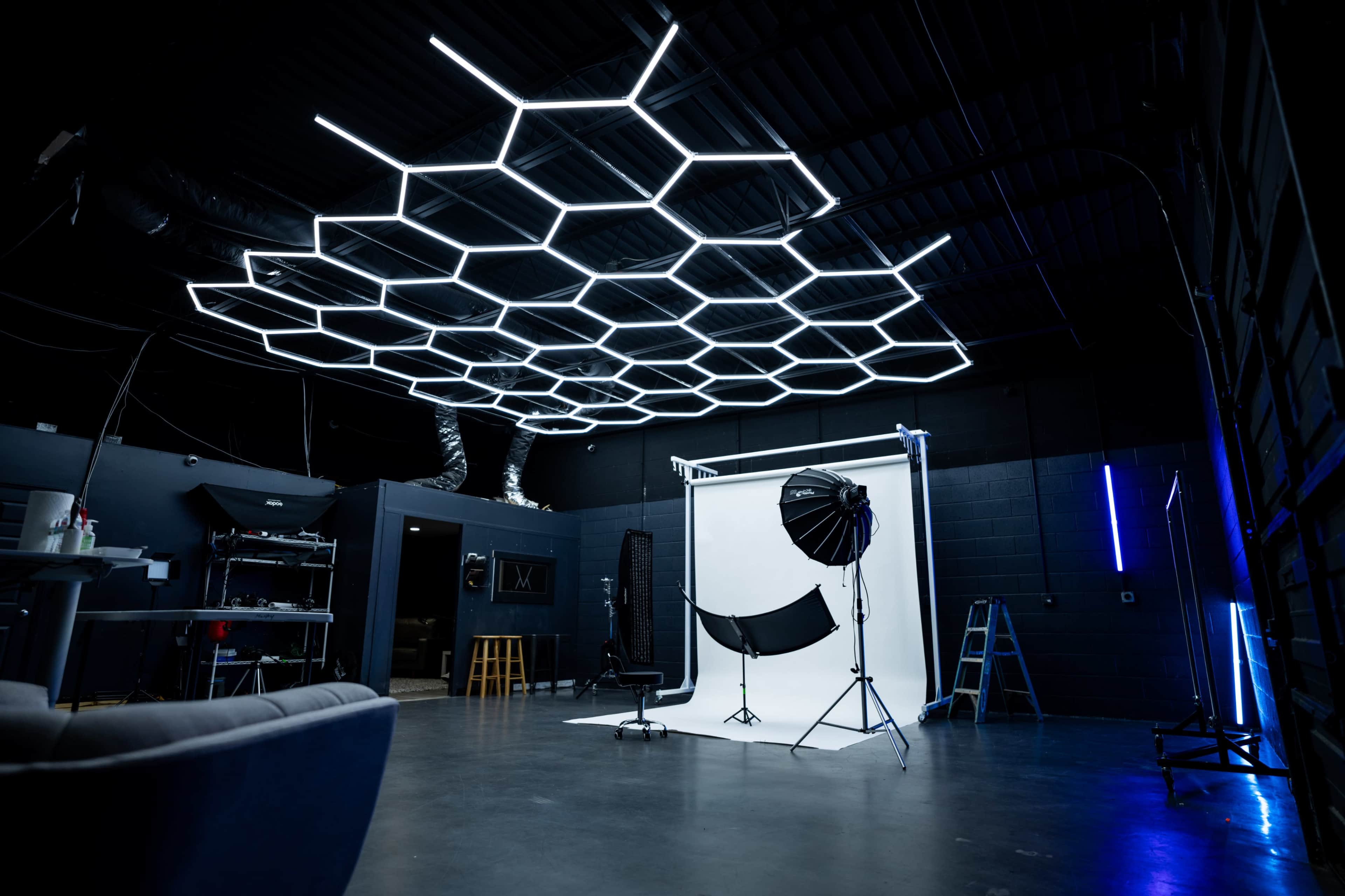 A photography studio with hexagonal LED lights overhead, a white backdrop setup, and various photography equipment arranged in the space.