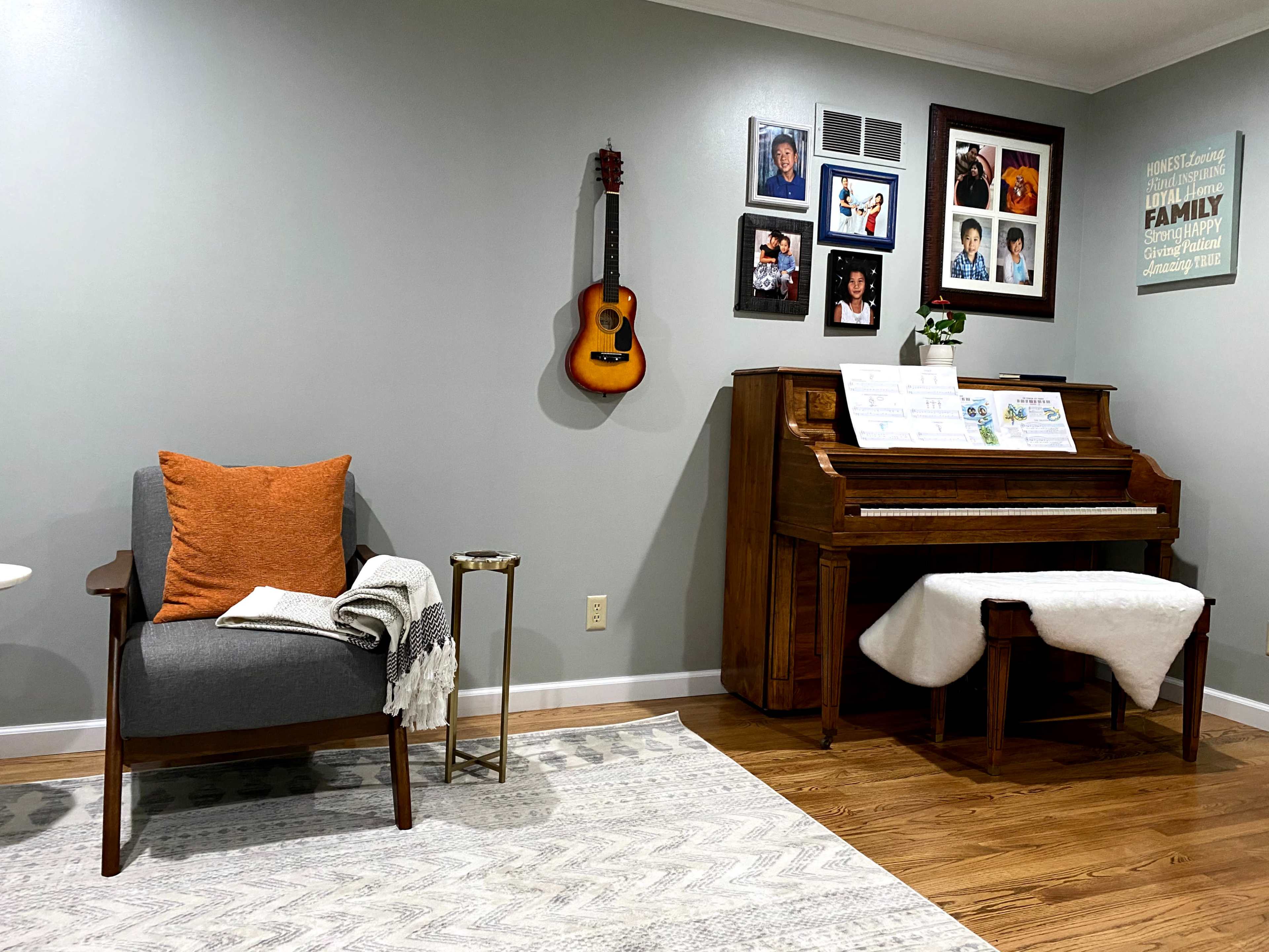 A cozy living room features a gray armchair with an orange pillow, an acoustic guitar and framed family photos on the wall, and a wooden piano with sheet music displayed.