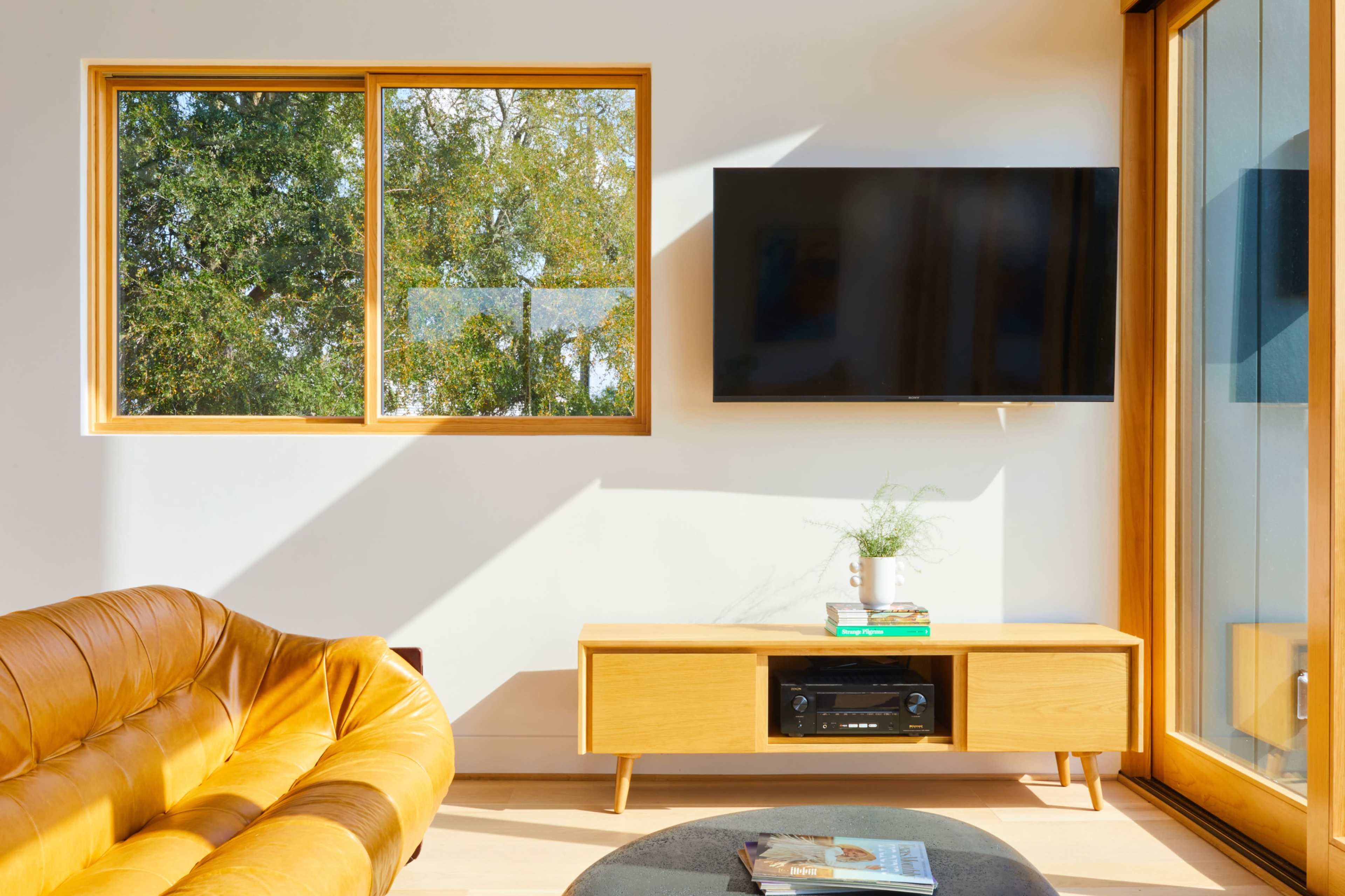 The image shows a modern living room with a leather sofa, a wooden entertainment unit with a television mounted on the wall, and a large window letting in natural light.
