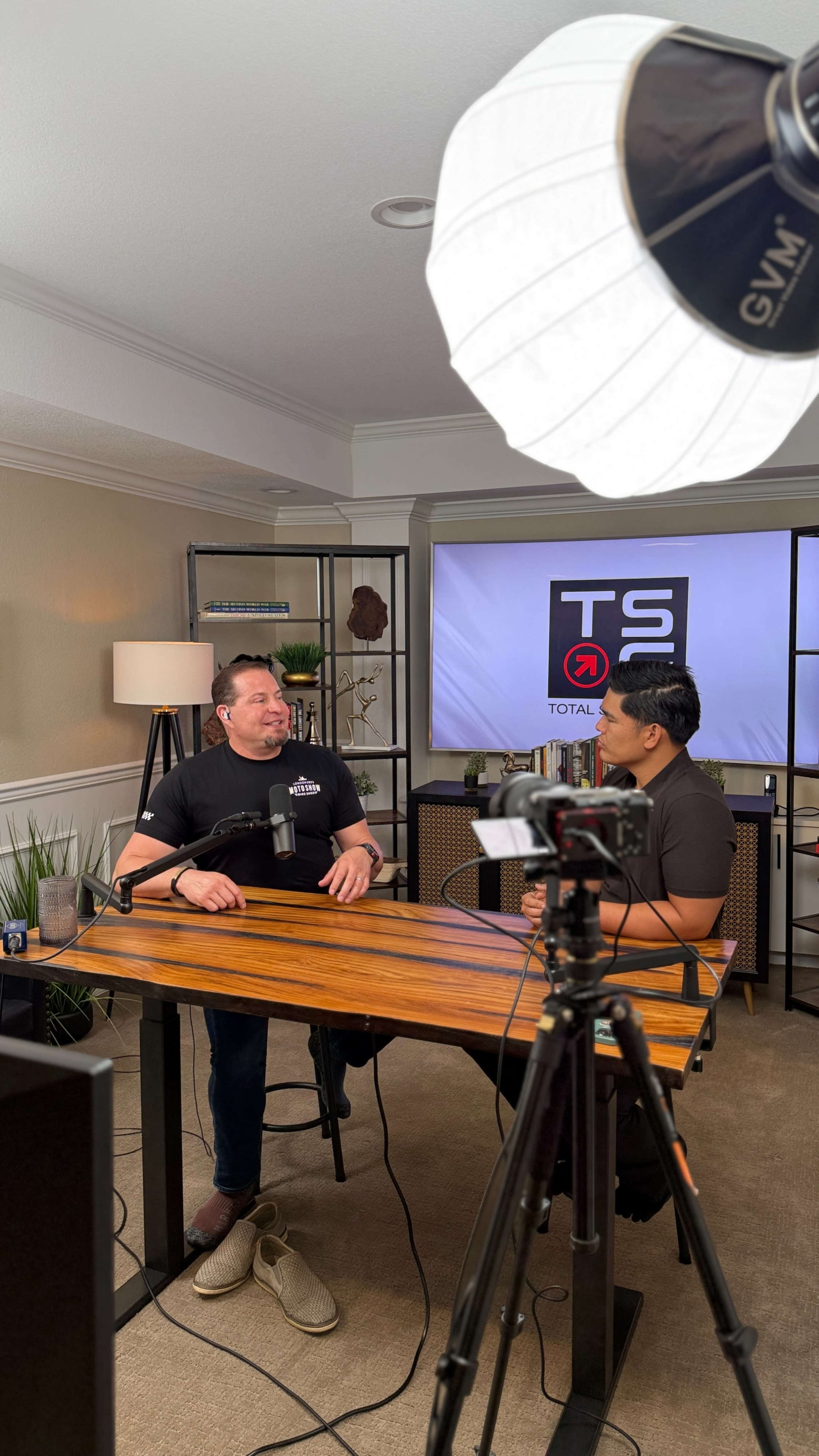 Two men are seated at a wooden table in a well-lit studio, discussing in front of a microphone and a camera, with a large display screen in the background showing the logo "TS" for Total Solutions.