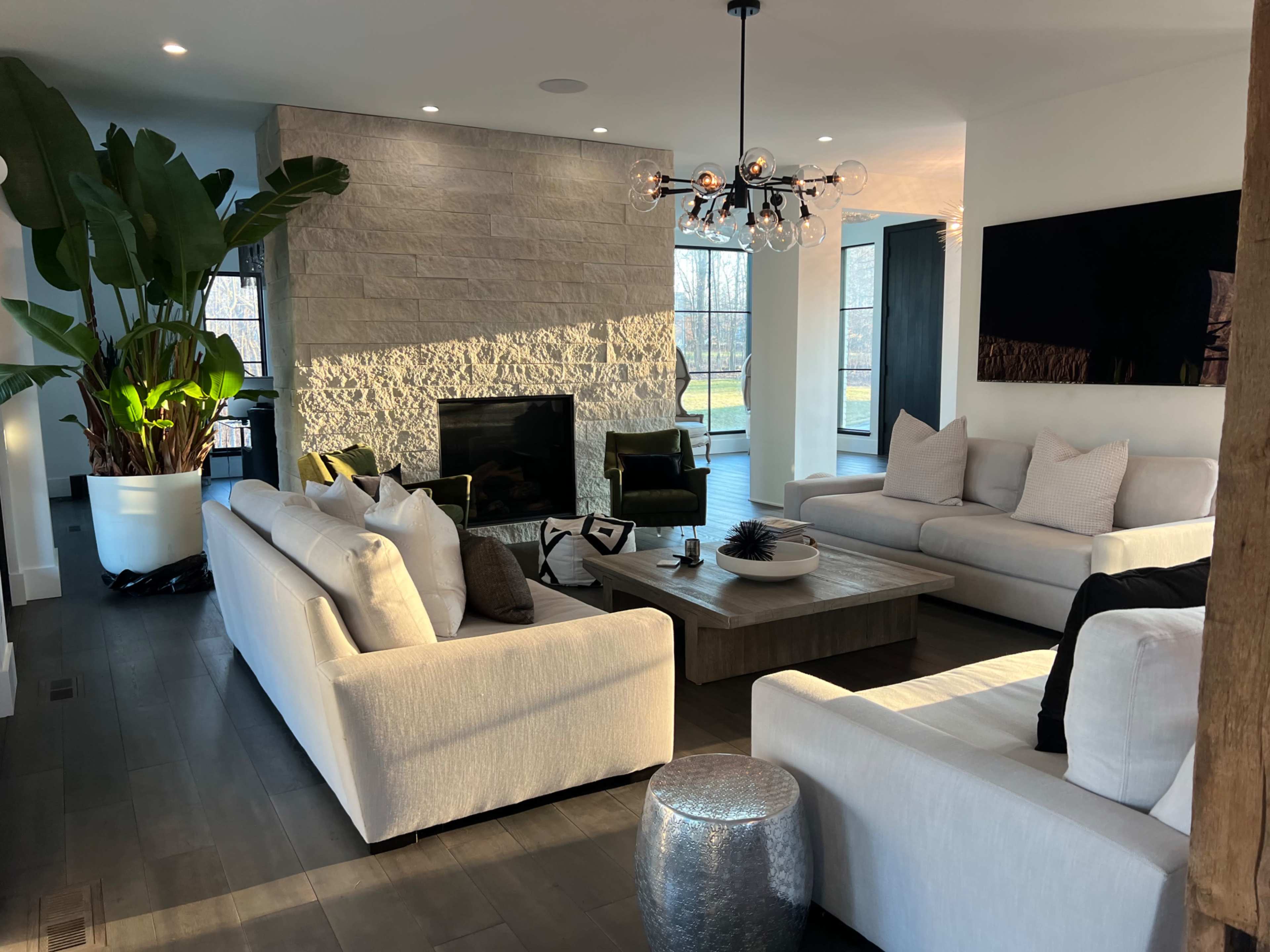 A modern living room featuring light-colored sofas, a stone fireplace, a large plant, and a coffee table in front of a window with natural light.