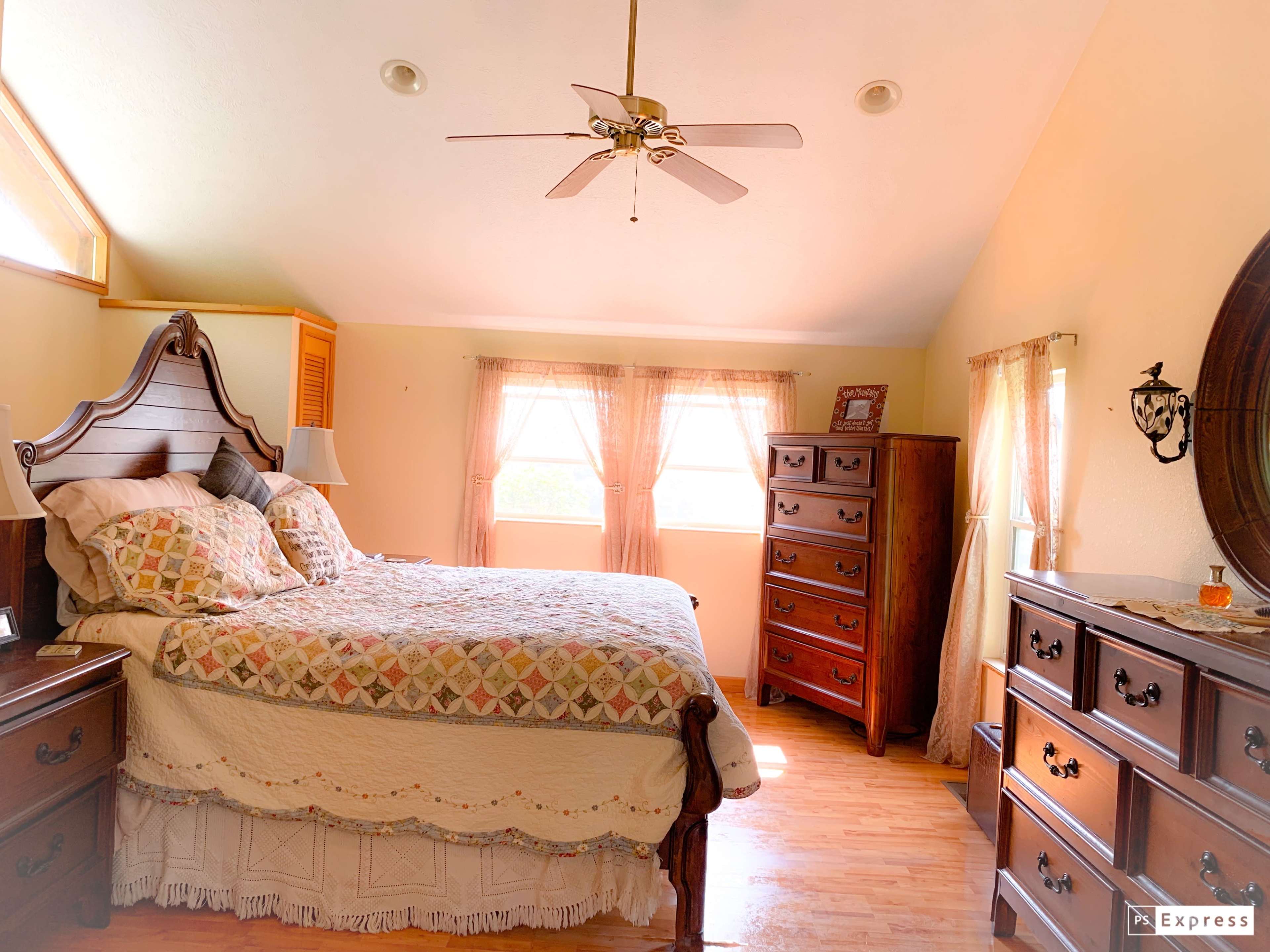 A well-furnished bedroom featuring a large bed, wooden dressers, and windows with light-colored curtains.