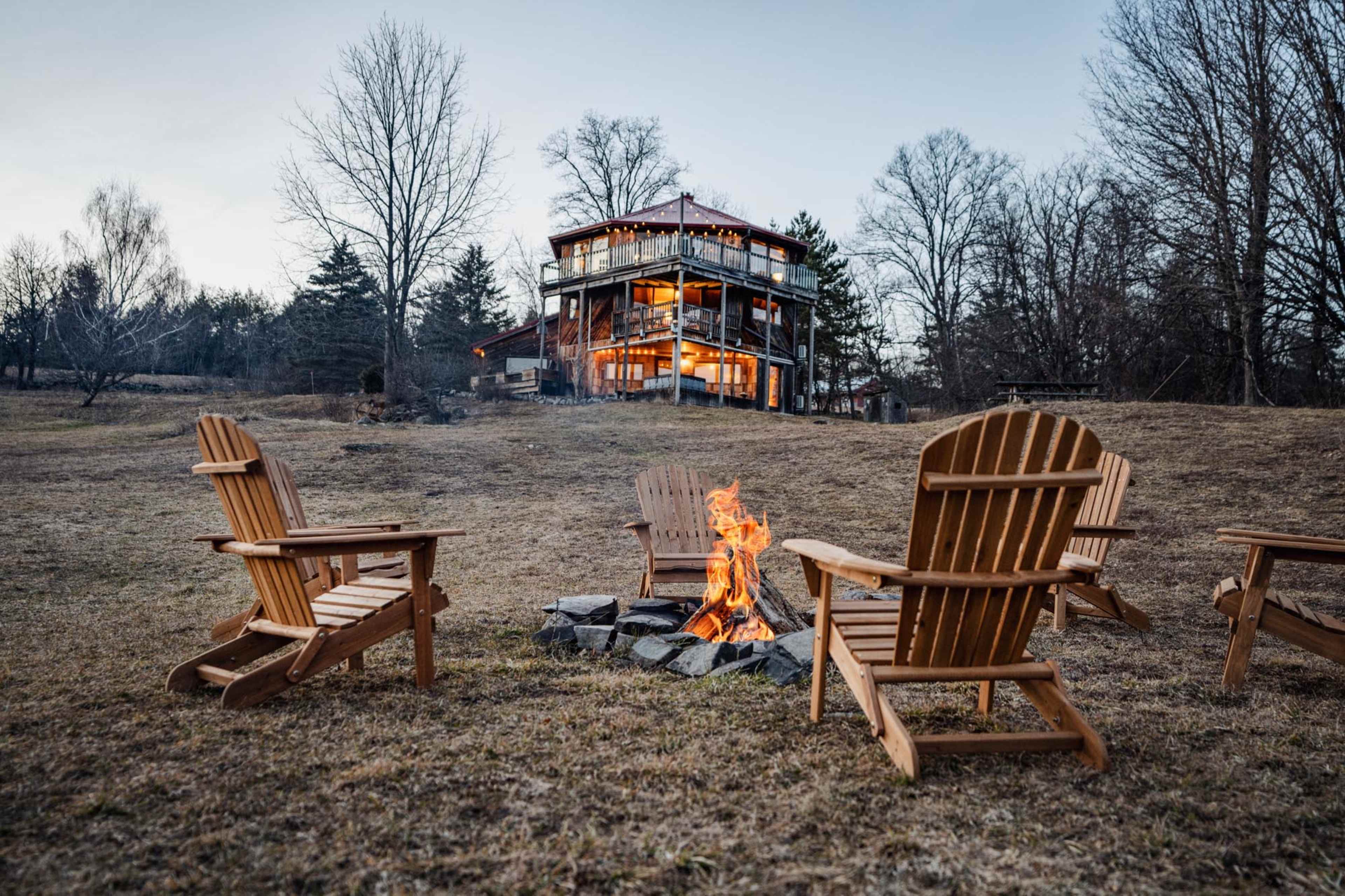 A circle of wooden chairs surrounds a fire pit with flames, while a multi-story building sits in the background against a twilight sky.