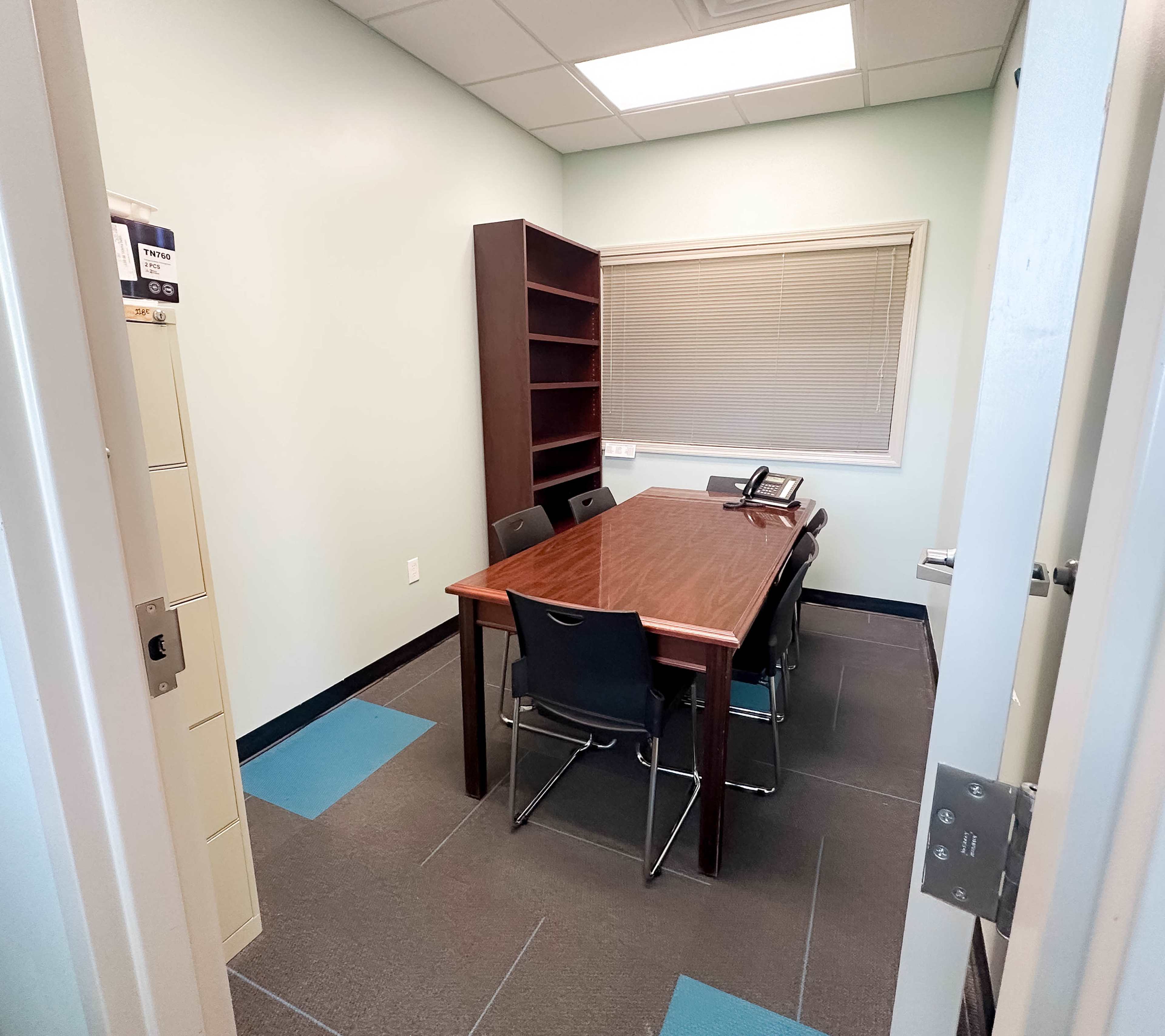 The image depicts a small meeting room with a wooden table surrounded by six chairs, a bookcase against one wall, and a phone on the table.