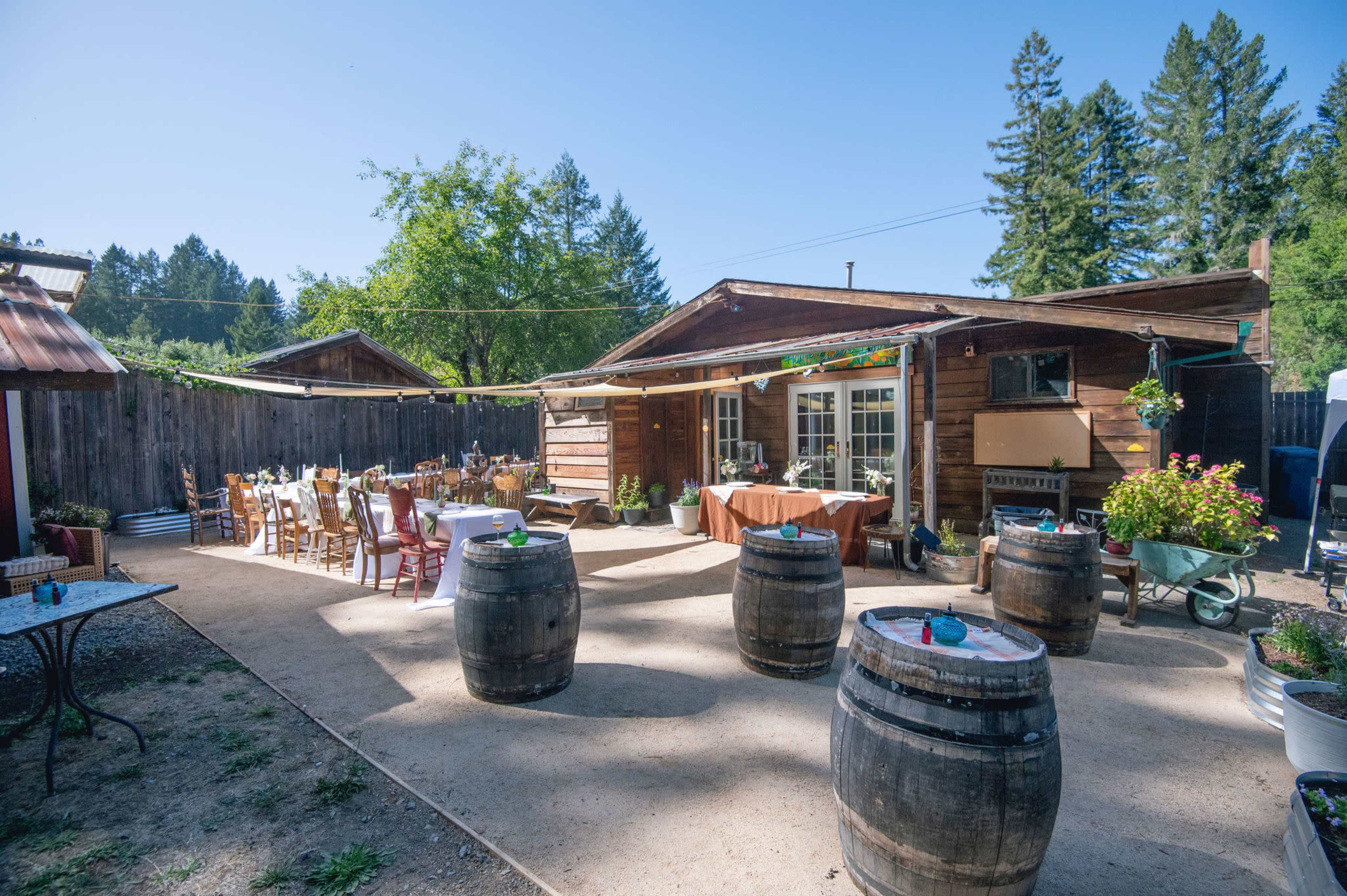 A rustic outdoor event space features wooden barrels, a dining area with tables and chairs, and a cabin surrounded by trees.