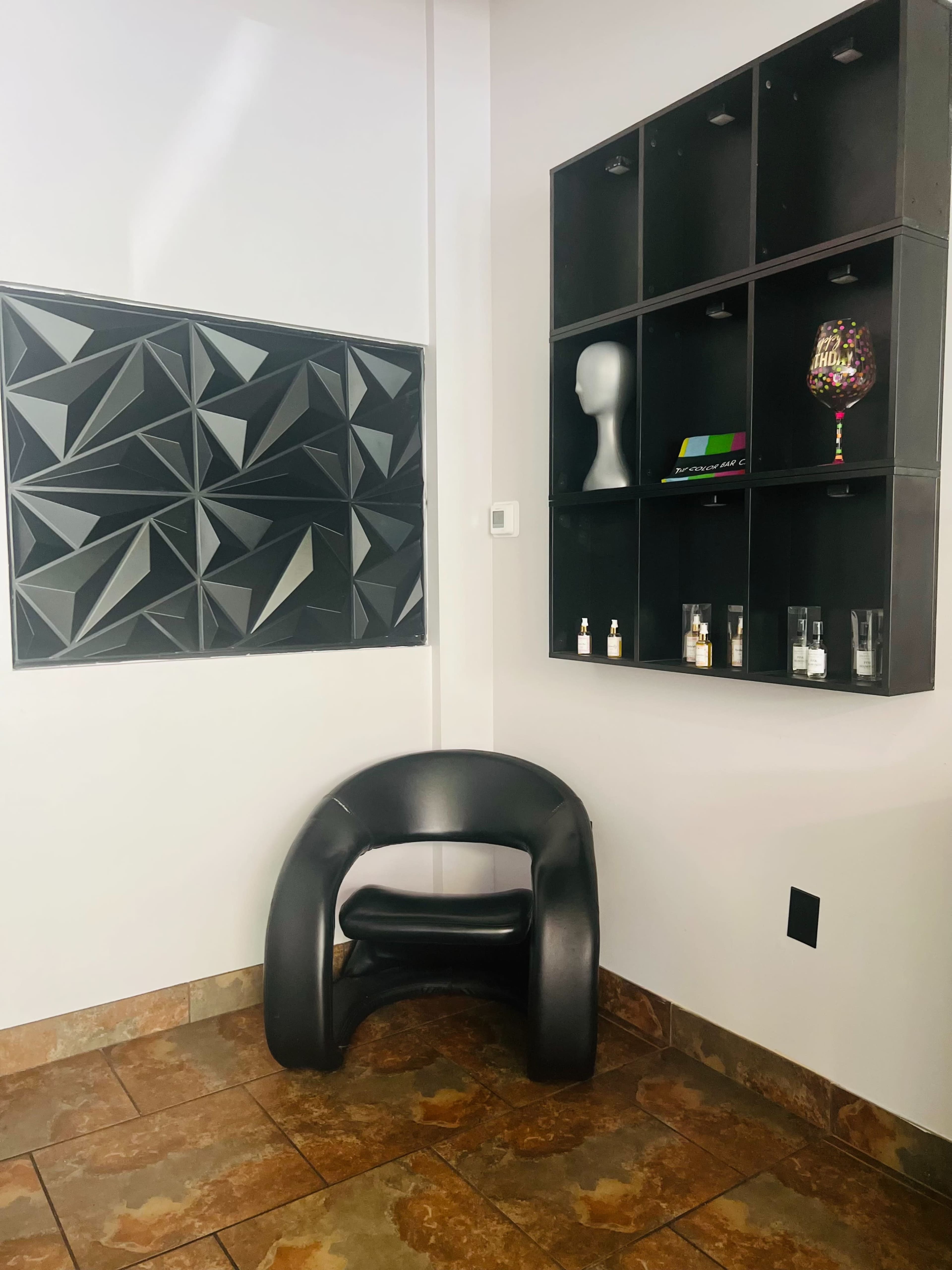 The image shows a corner of a room featuring a black geometric wall panel, a black chair, and a shelf with various small items.