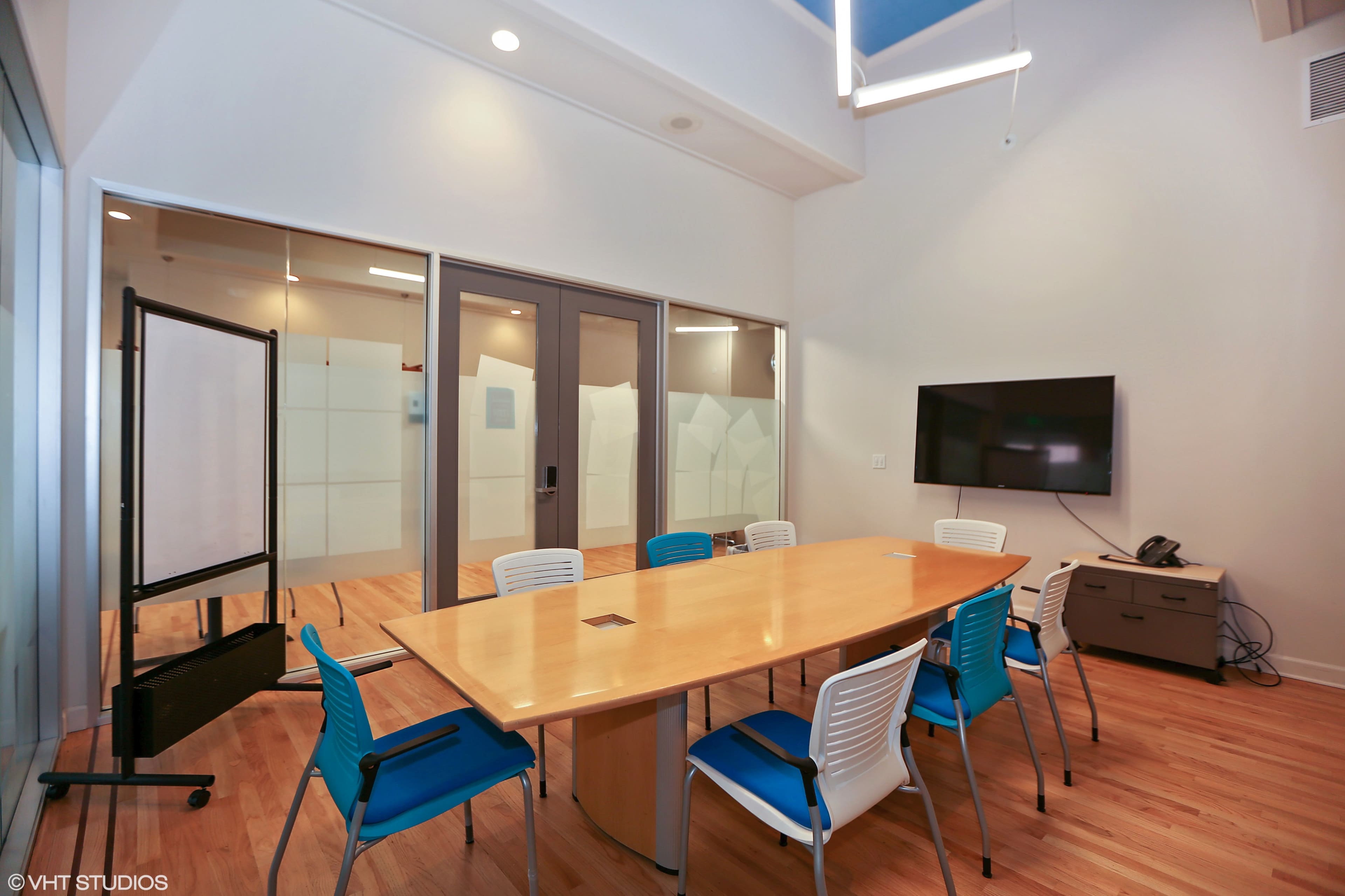 The image shows a conference room featuring a long wooden table surrounded by white and teal chairs, with a wall-mounted TV and a nearby glass wall.
