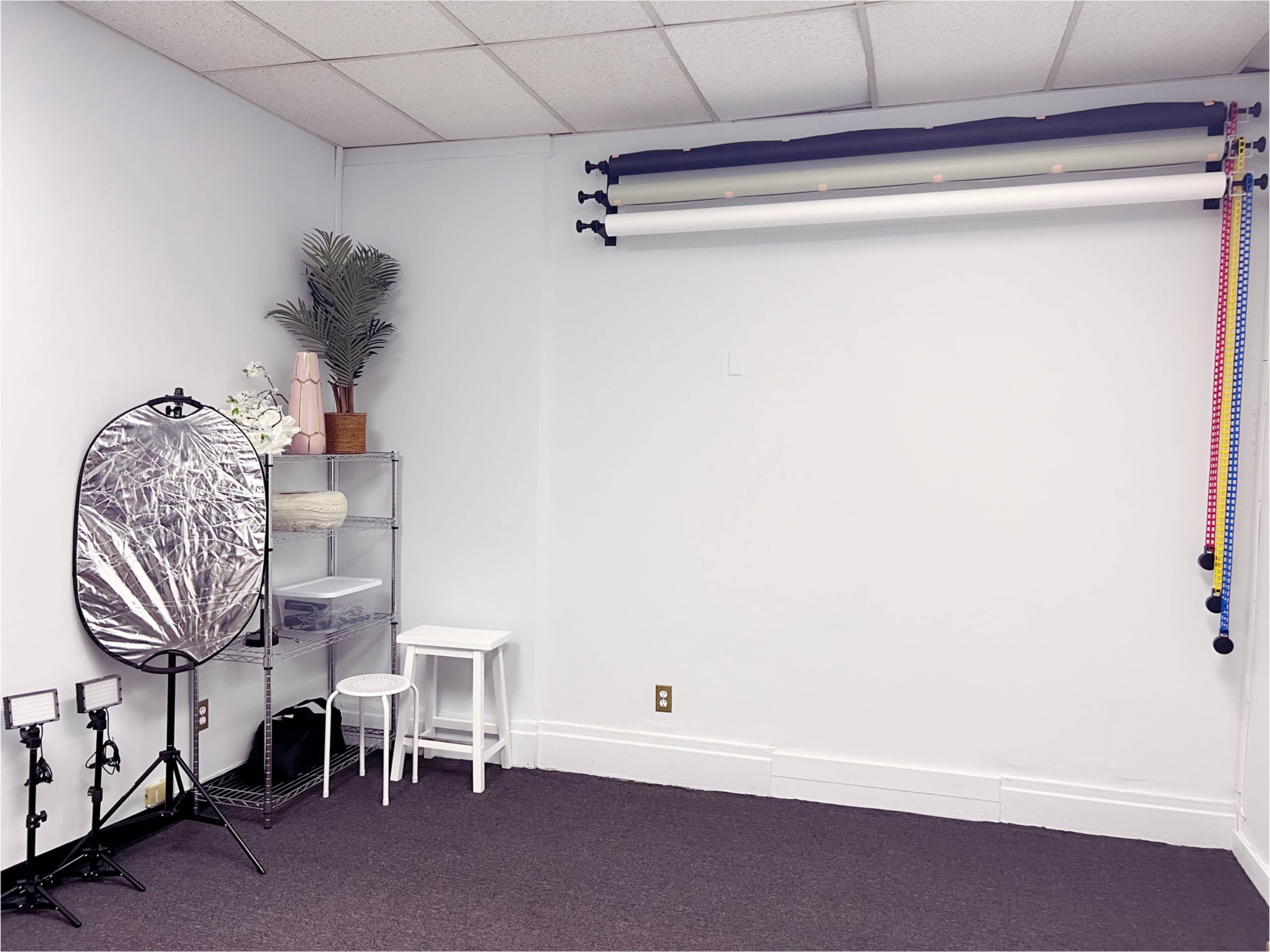 The image shows a minimalist studio space with a white wall, a rack of backdrop rolls, a reflective circular disc on a stand, a clear storage unit, a small white stool, and several light stands.