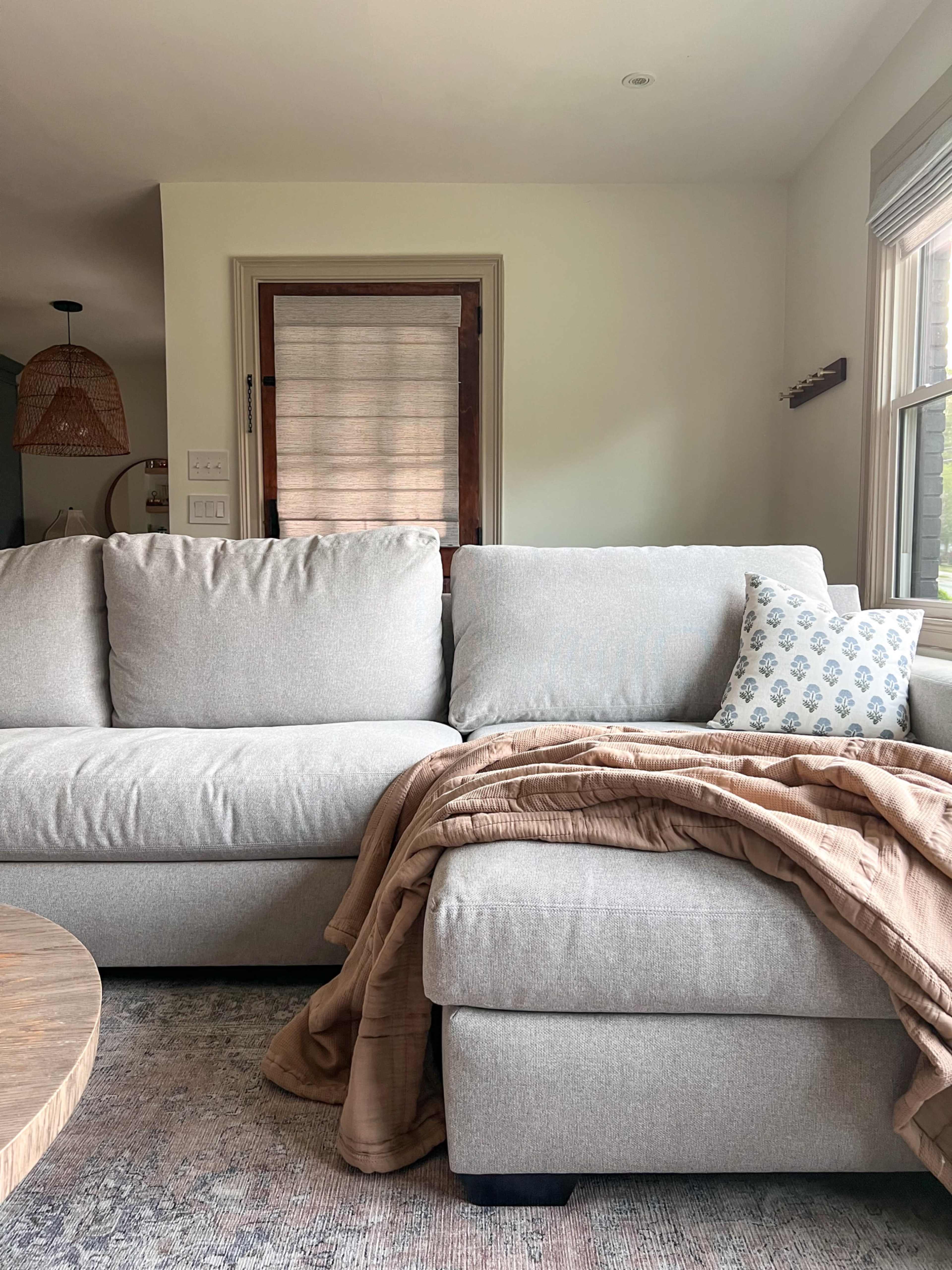A light gray sectional sofa with a patterned pillow and a beige throw blanket is positioned near a window with natural light streaming in.