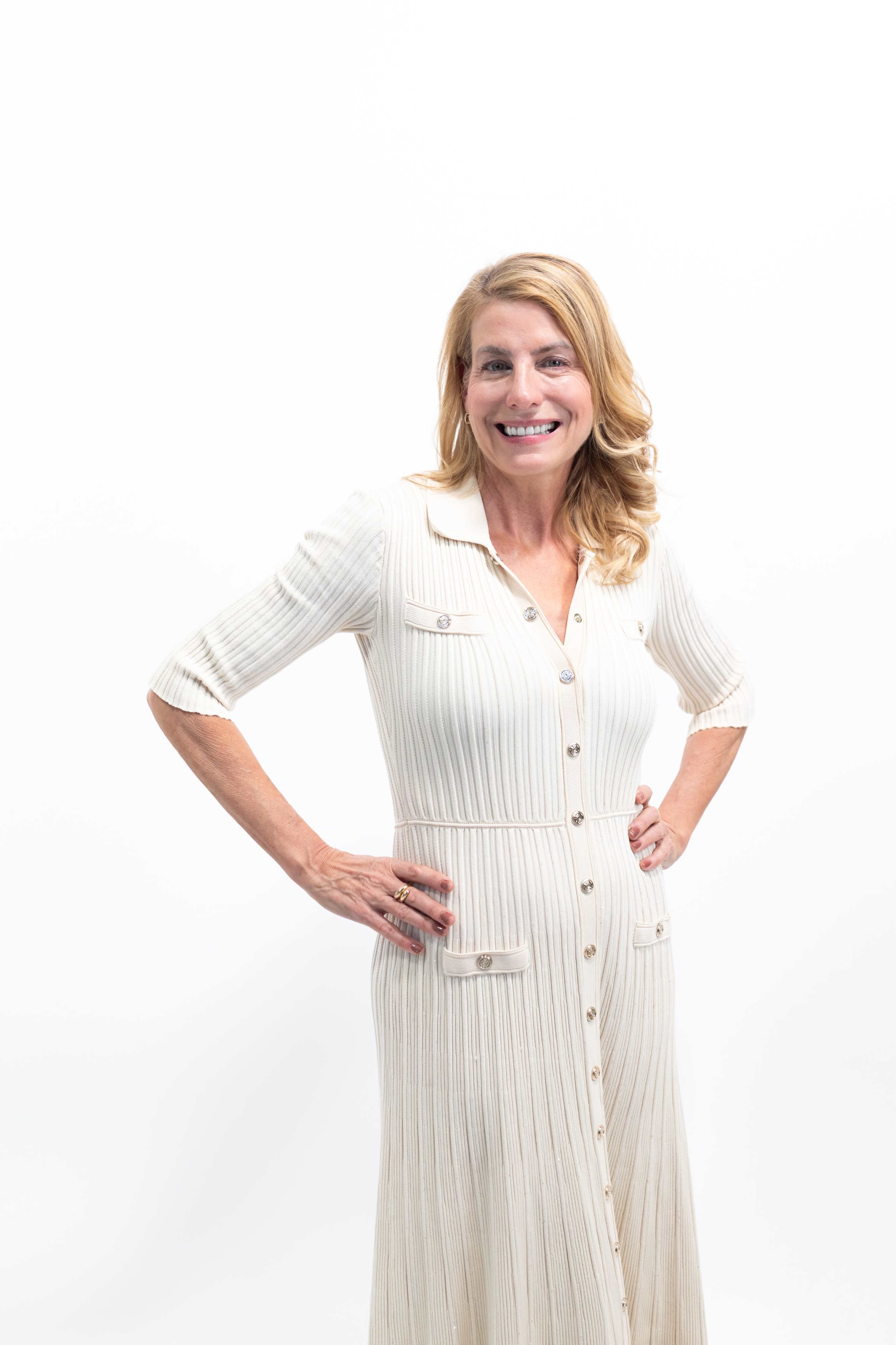 A woman stands with her hands on her hips, wearing a light-colored, ribbed dress and smiling against a white background.