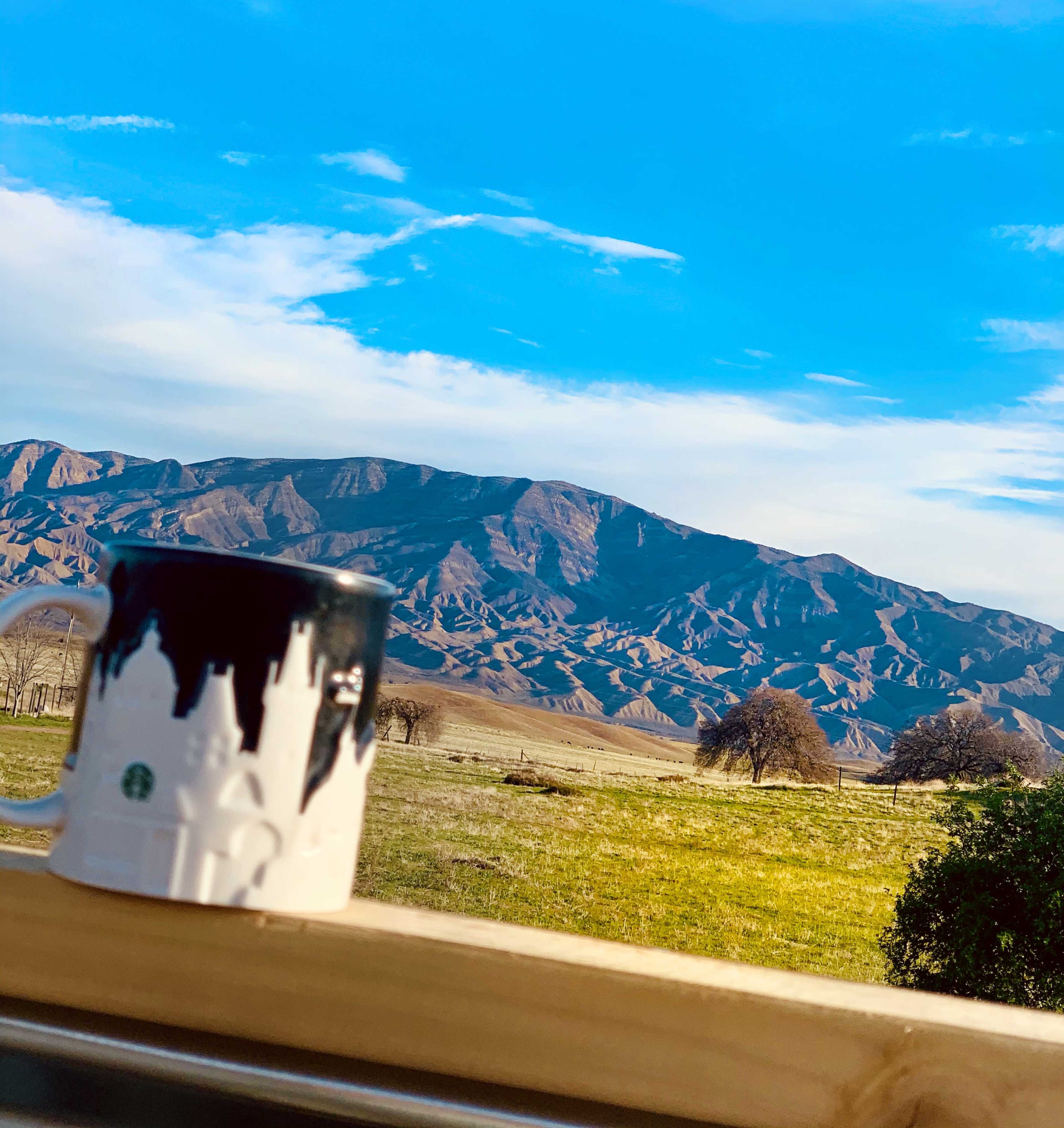 A coffee mug is placed on a wooden railing in the foreground, overlooking a mountainous landscape under a clear blue sky.