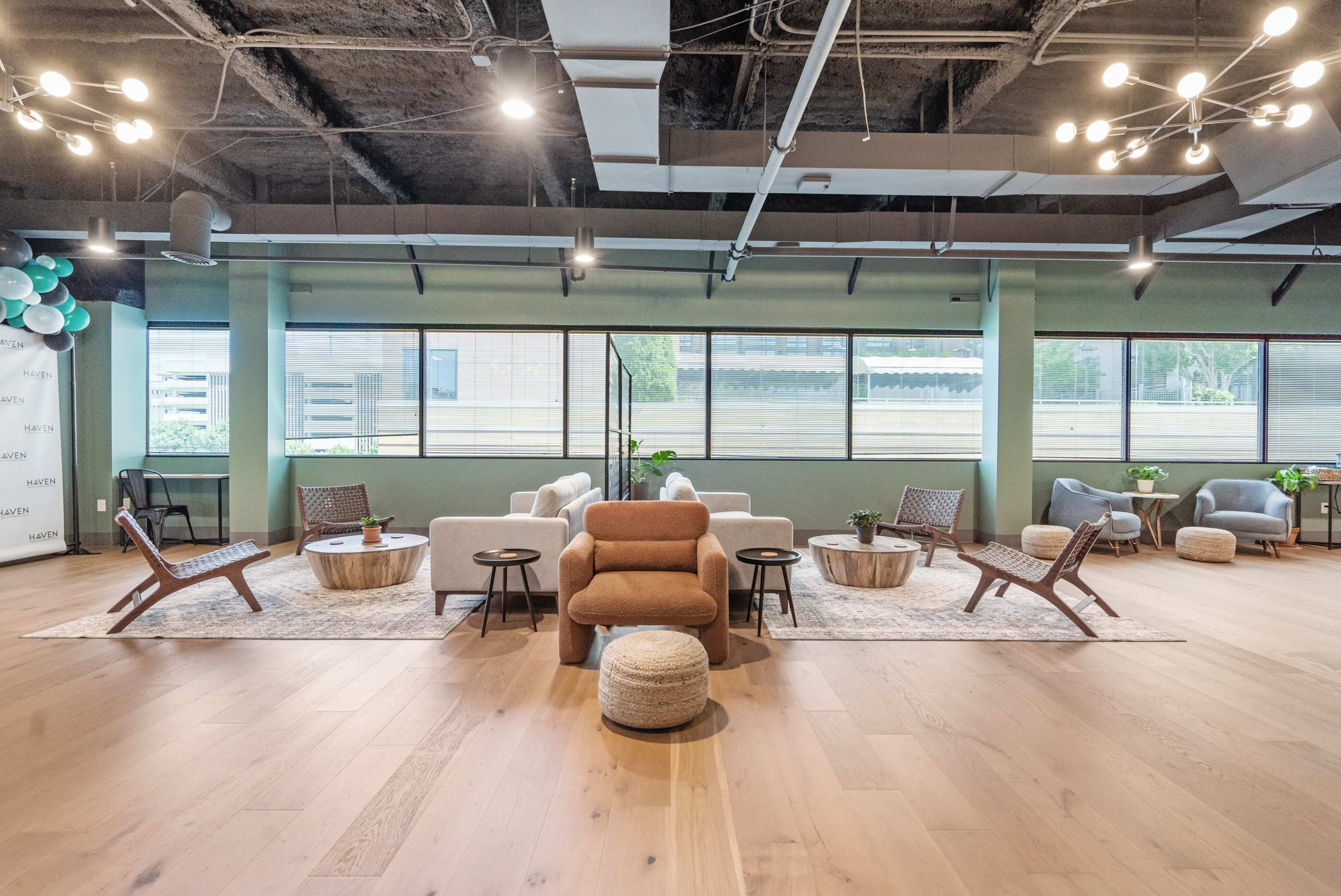 The image shows a modern, open lounge area with several seating arrangements, including sofas and armchairs, surrounded by large windows allowing natural light in.
