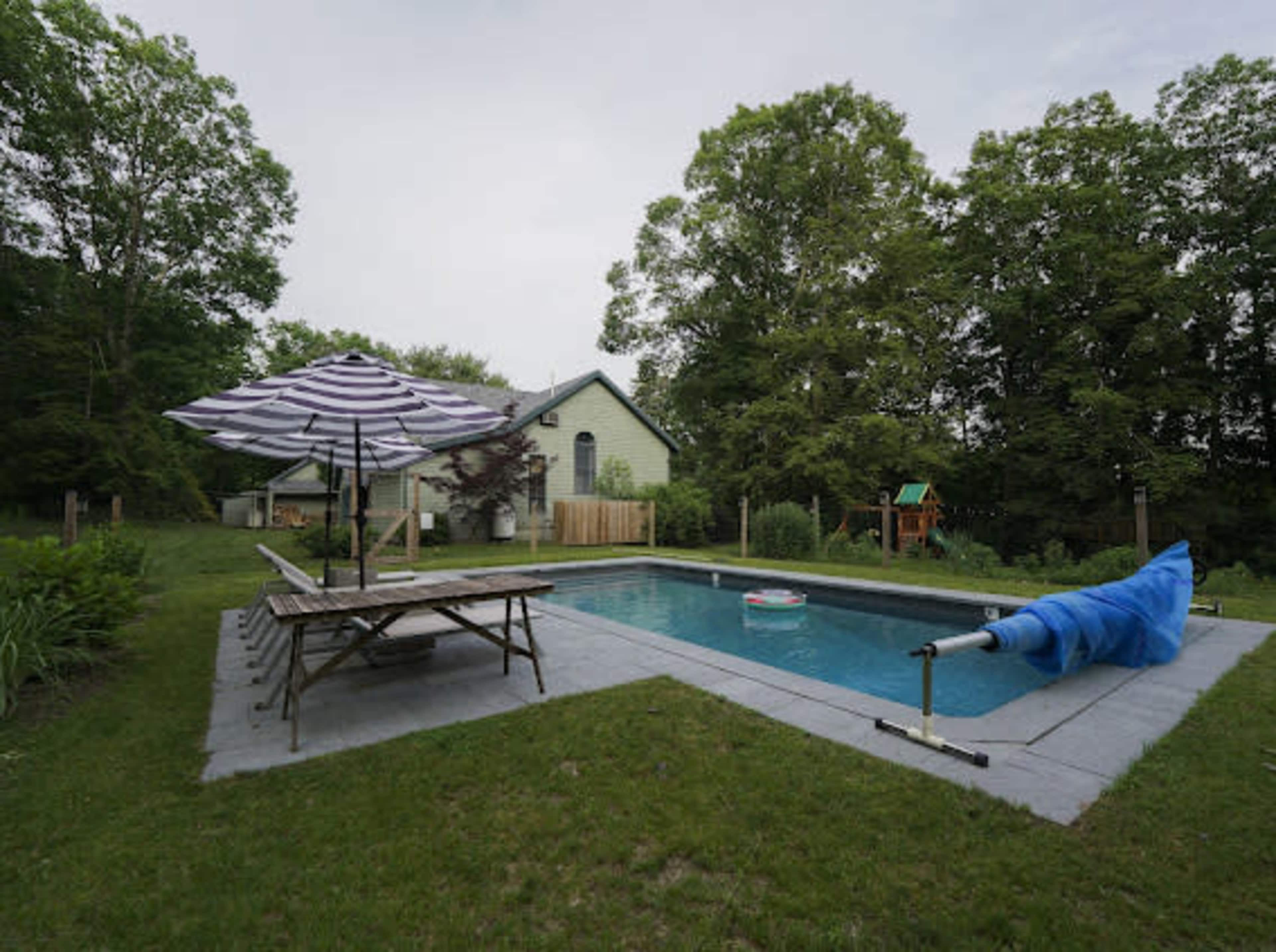 The image shows a backyard with a swimming pool surrounded by grass, a picnic table, an umbrella, and a house in the background.