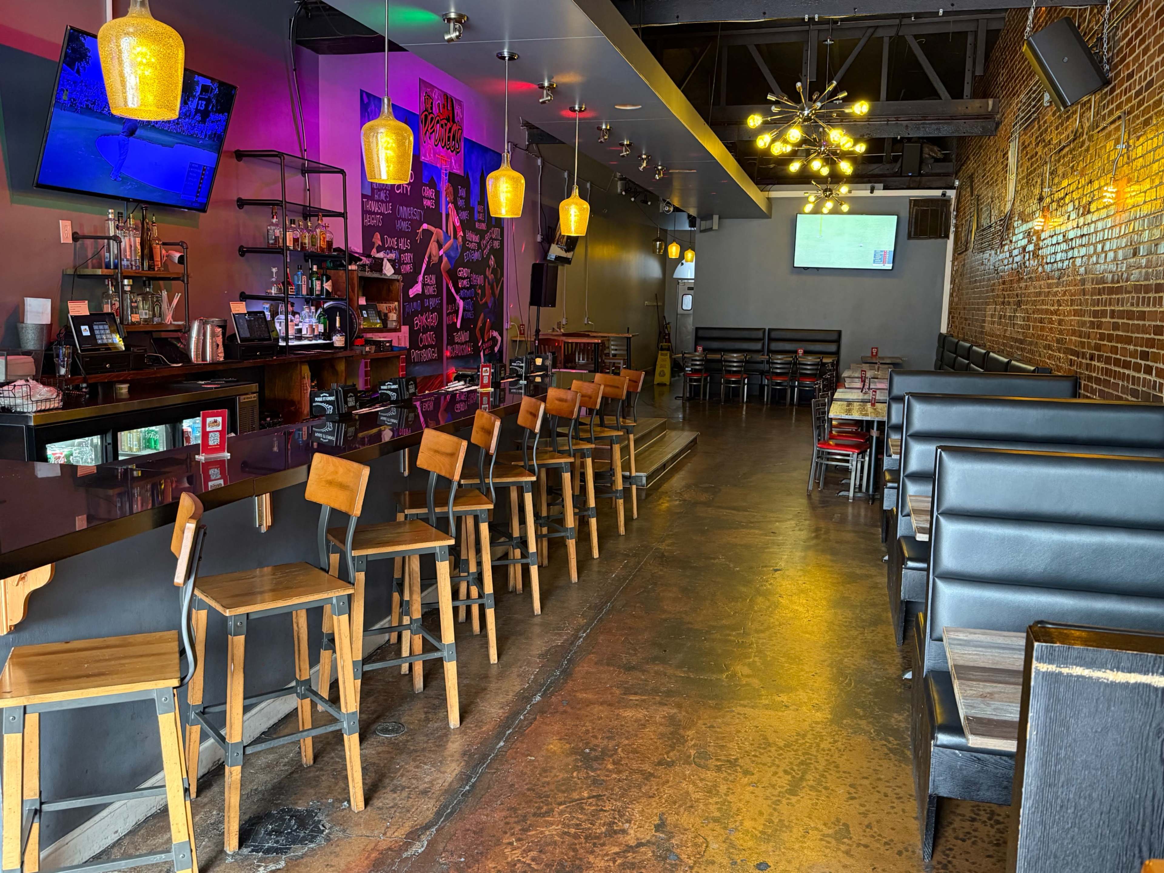The image shows a modern bar and dining area with wooden seating, colorful lighting, and a long counter, featuring a television mounted on the wall.