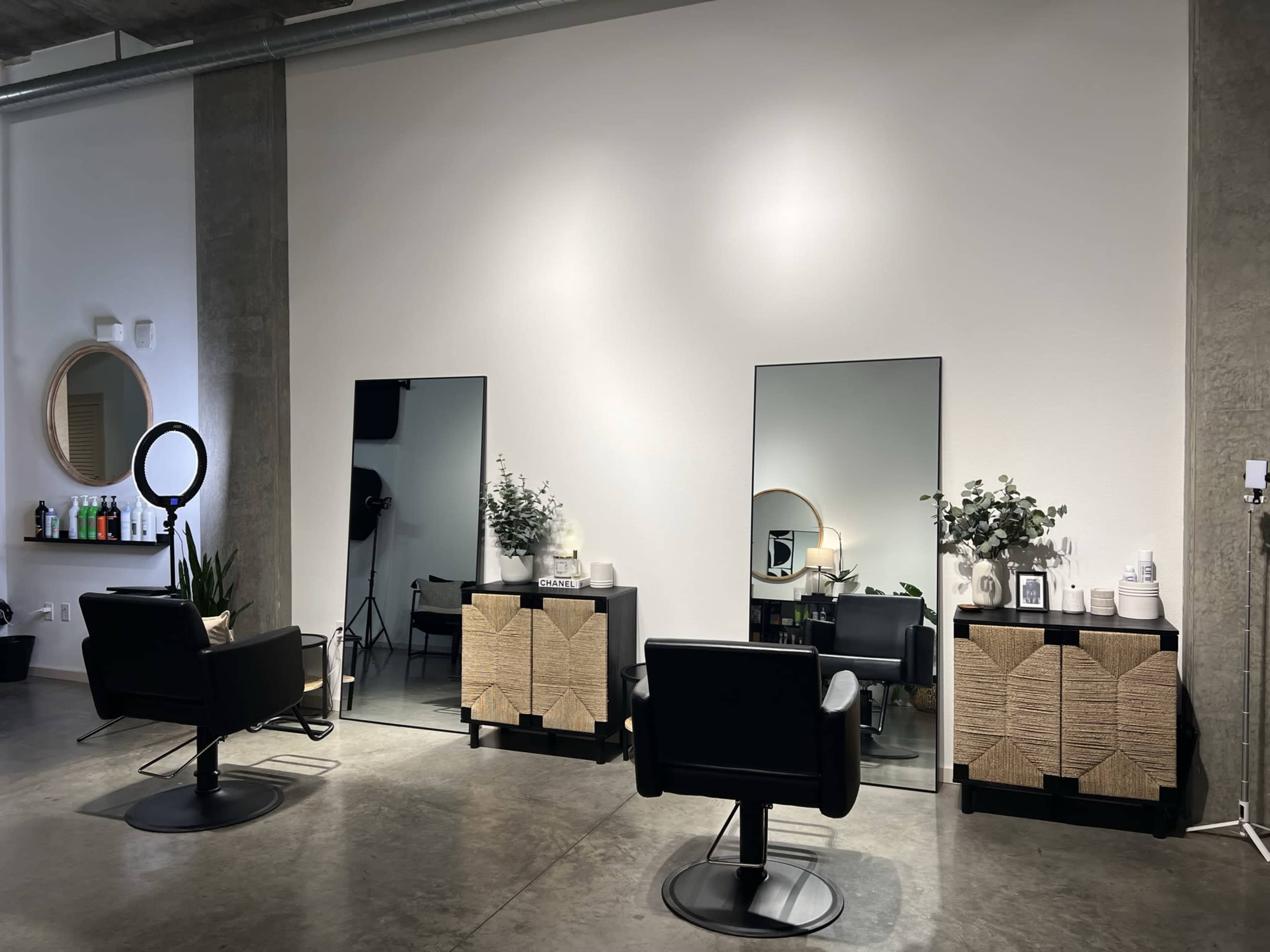 The image shows a modern hair salon featuring two styling stations with mirrors, black chairs, and decorative elements on wooden cabinets against a neutral wall.