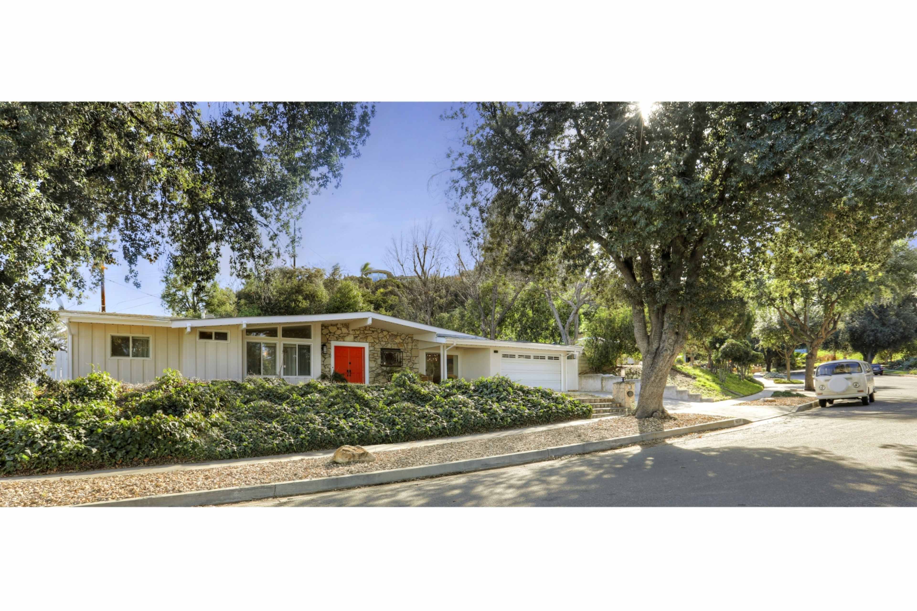 A single-story house with a red front door is surrounded by lush greenery on a quiet street.