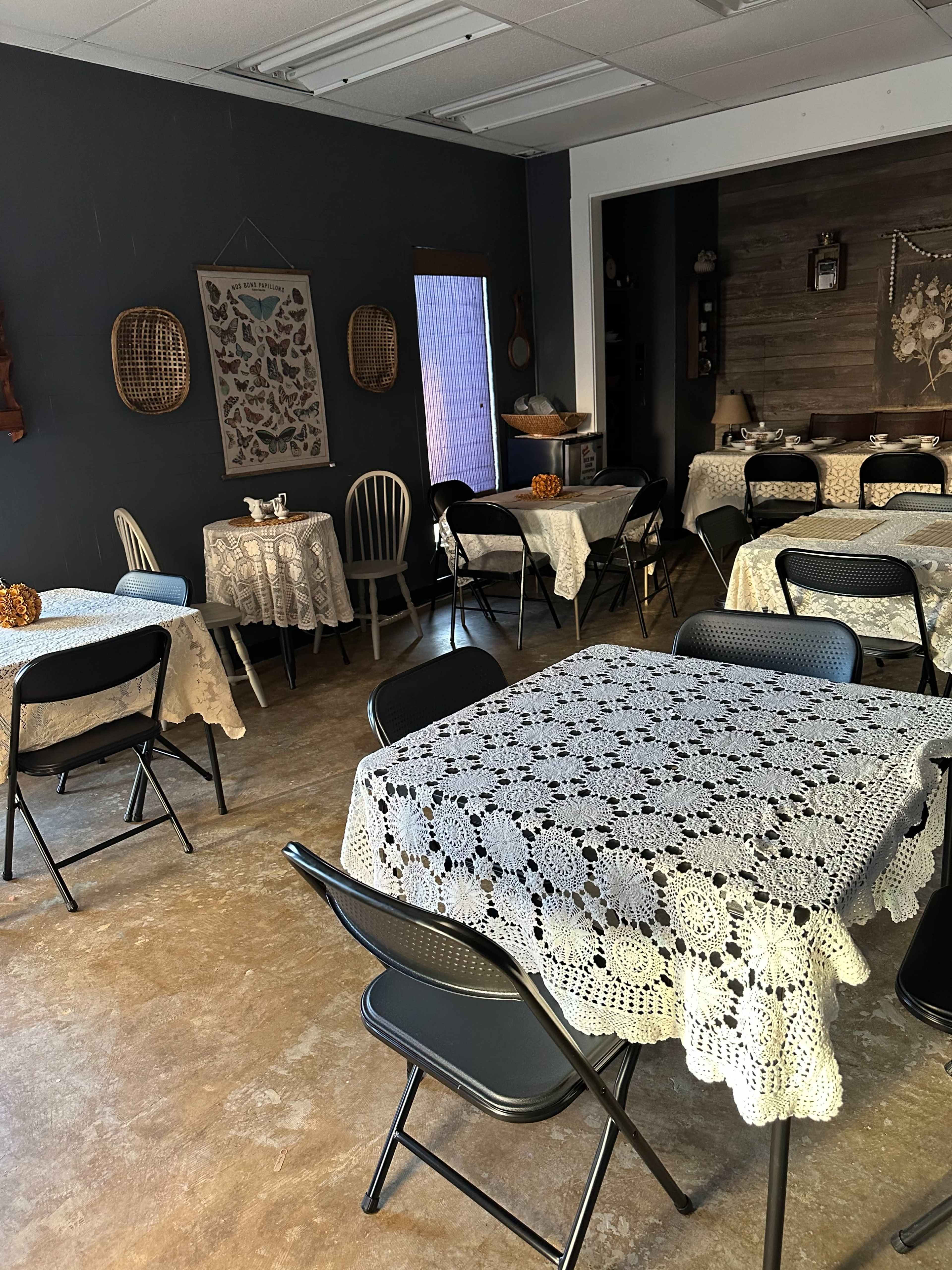 A café interior with several tables covered in lace tablecloths and simple black chairs arranged throughout the space.