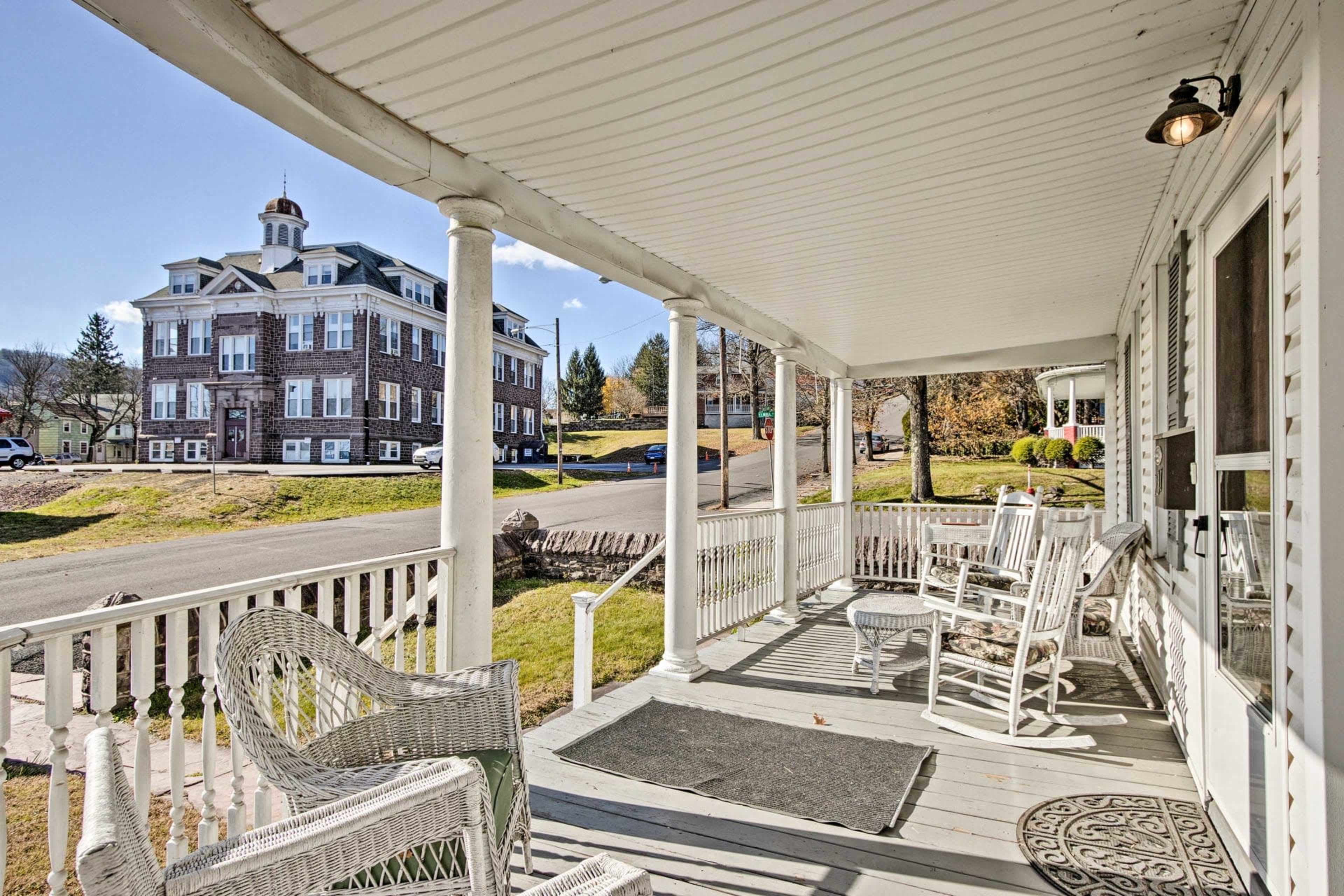 A porch with white rocking chairs, overlooking a street and a large building in the background.