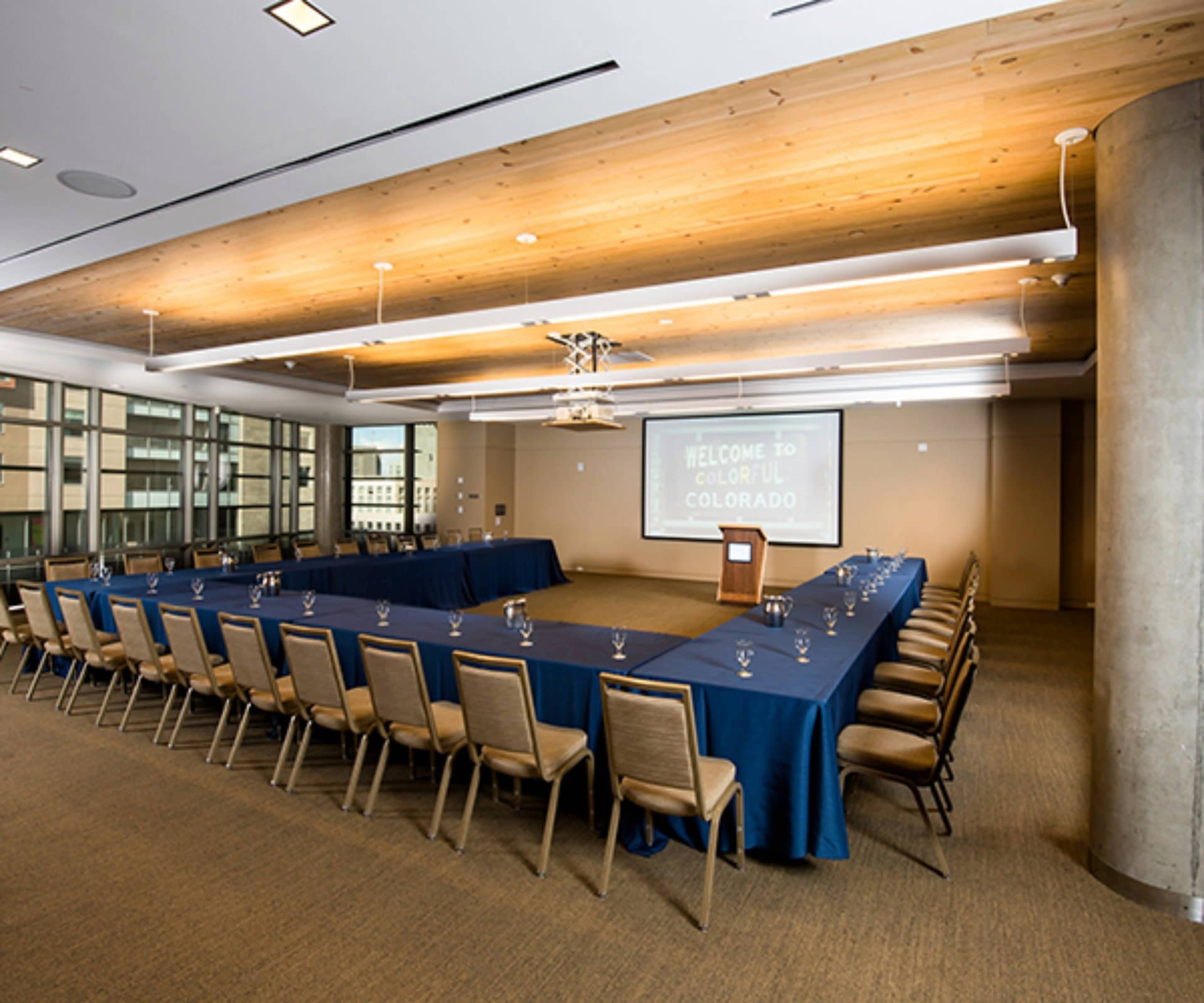 A large conference room arranged in a U-shape with a podium at one end and a screen displaying a welcome message in the background.