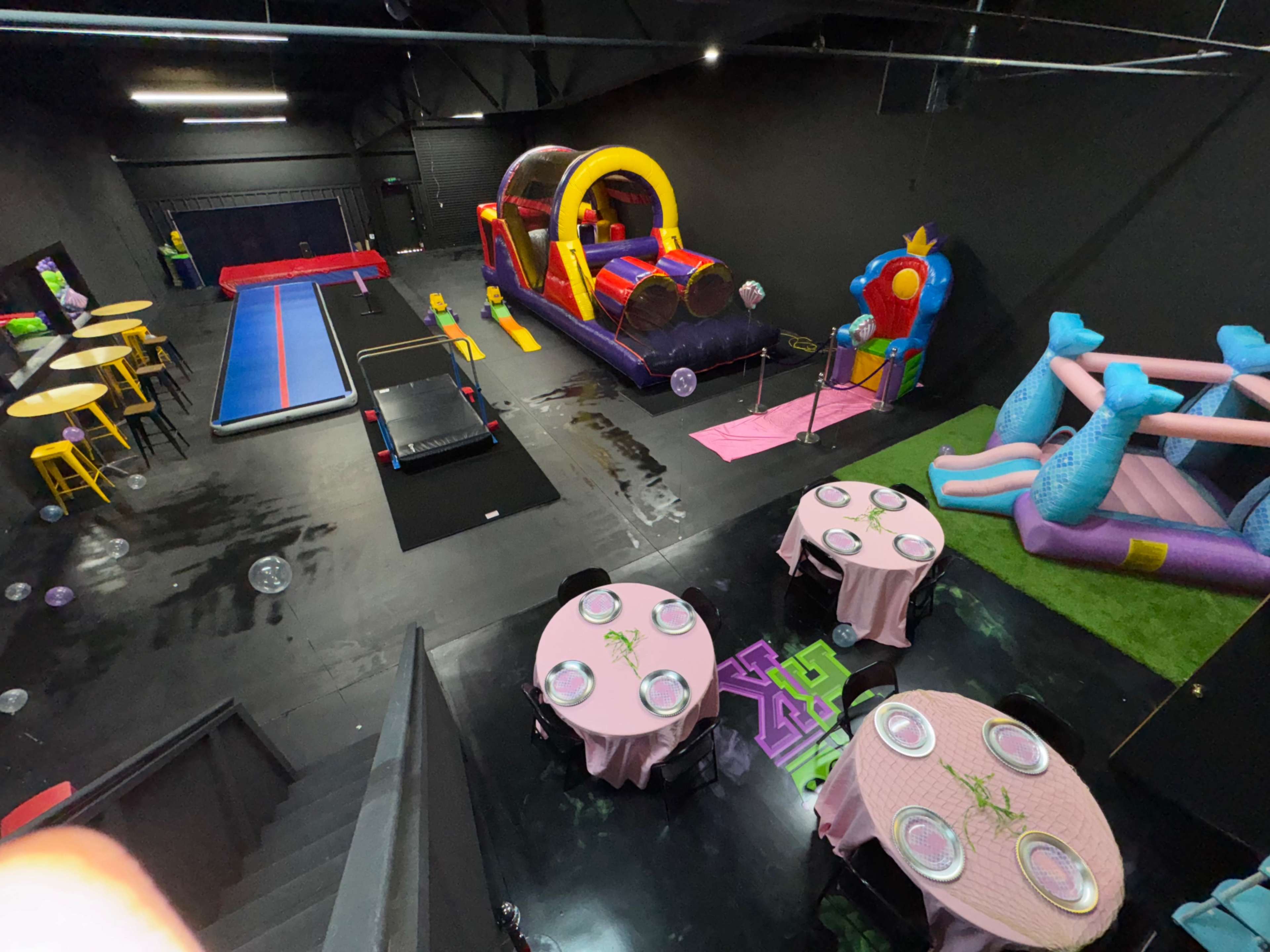 The image shows a large indoor space with inflatable play structures, a bounce house, and tables set for an event.