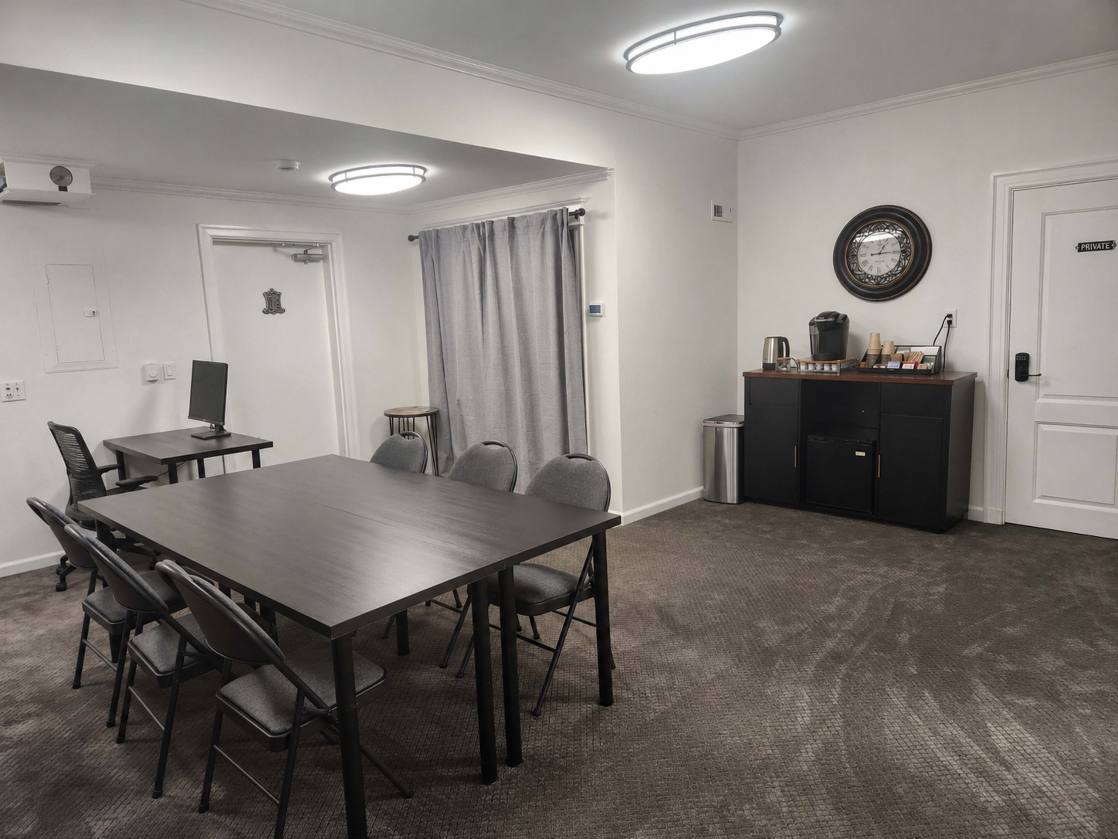 The image shows a conference room with a rectangular table surrounded by six chairs, a small kitchenette area, and a door with a number.
