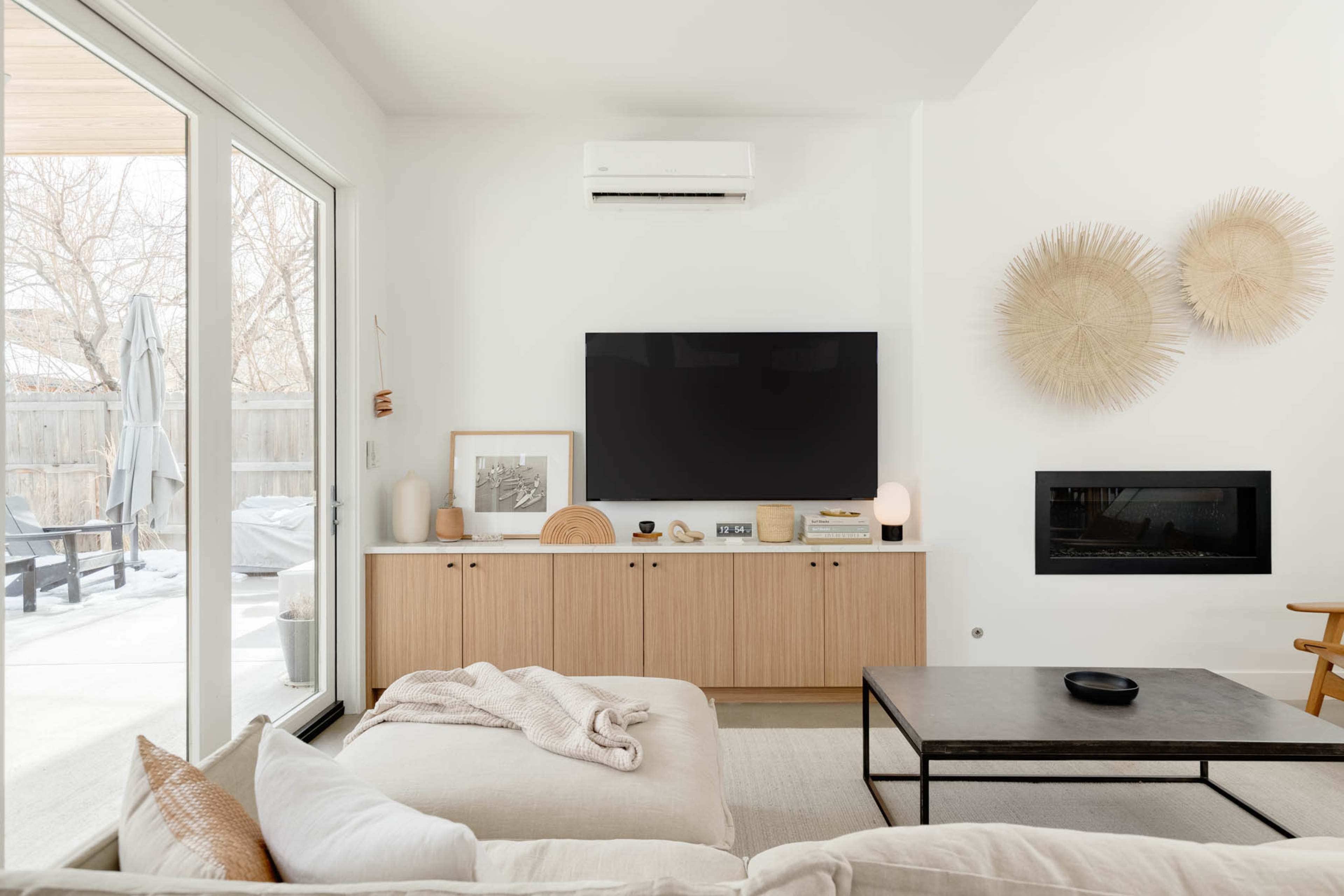 A modern living room features a large flat-screen TV mounted on a wall above a light wood cabinet, with a coffee table in front and sliding glass doors leading to an outdoor space.