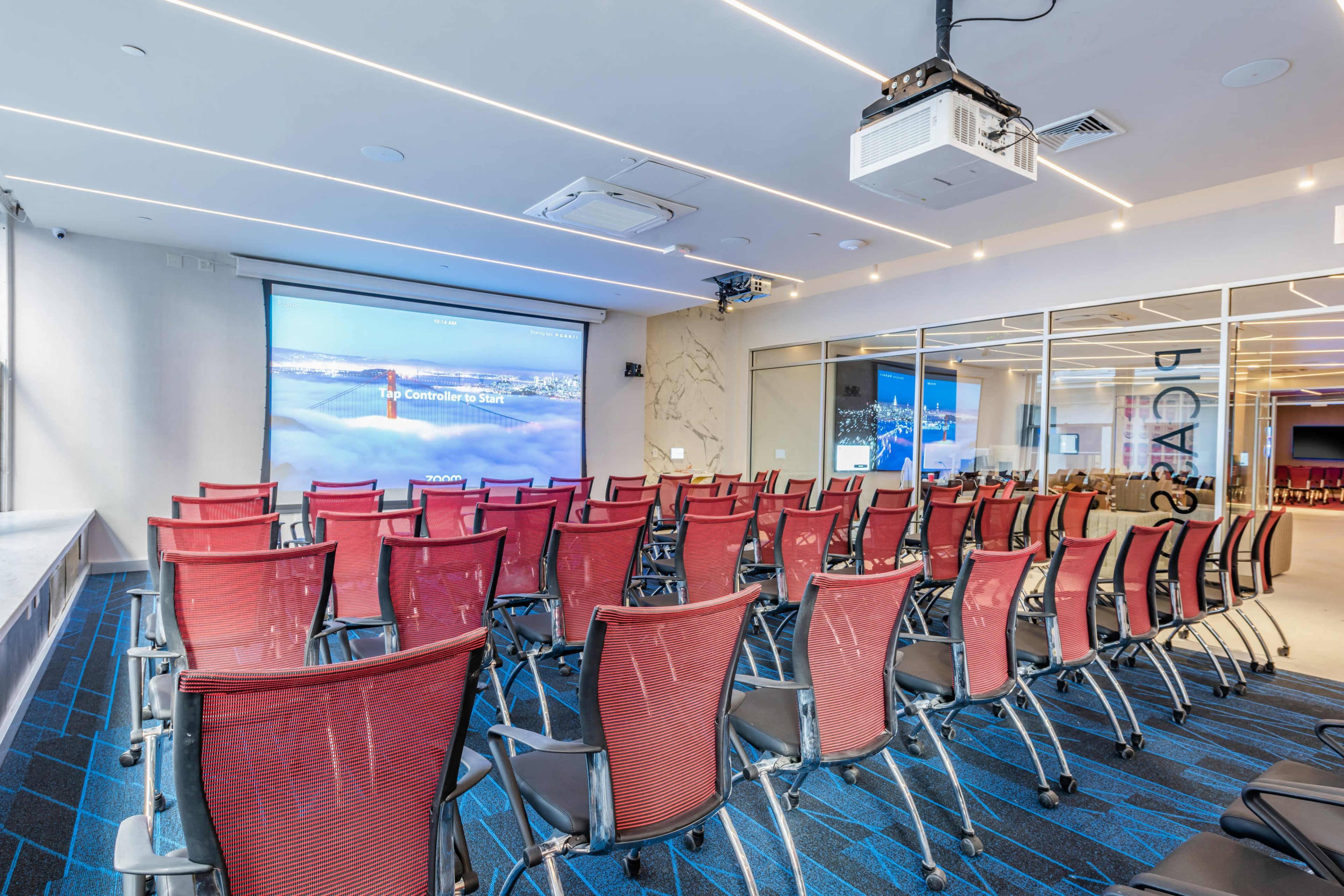 The image shows a modern conference room with red chairs arranged in rows facing a projection screen.