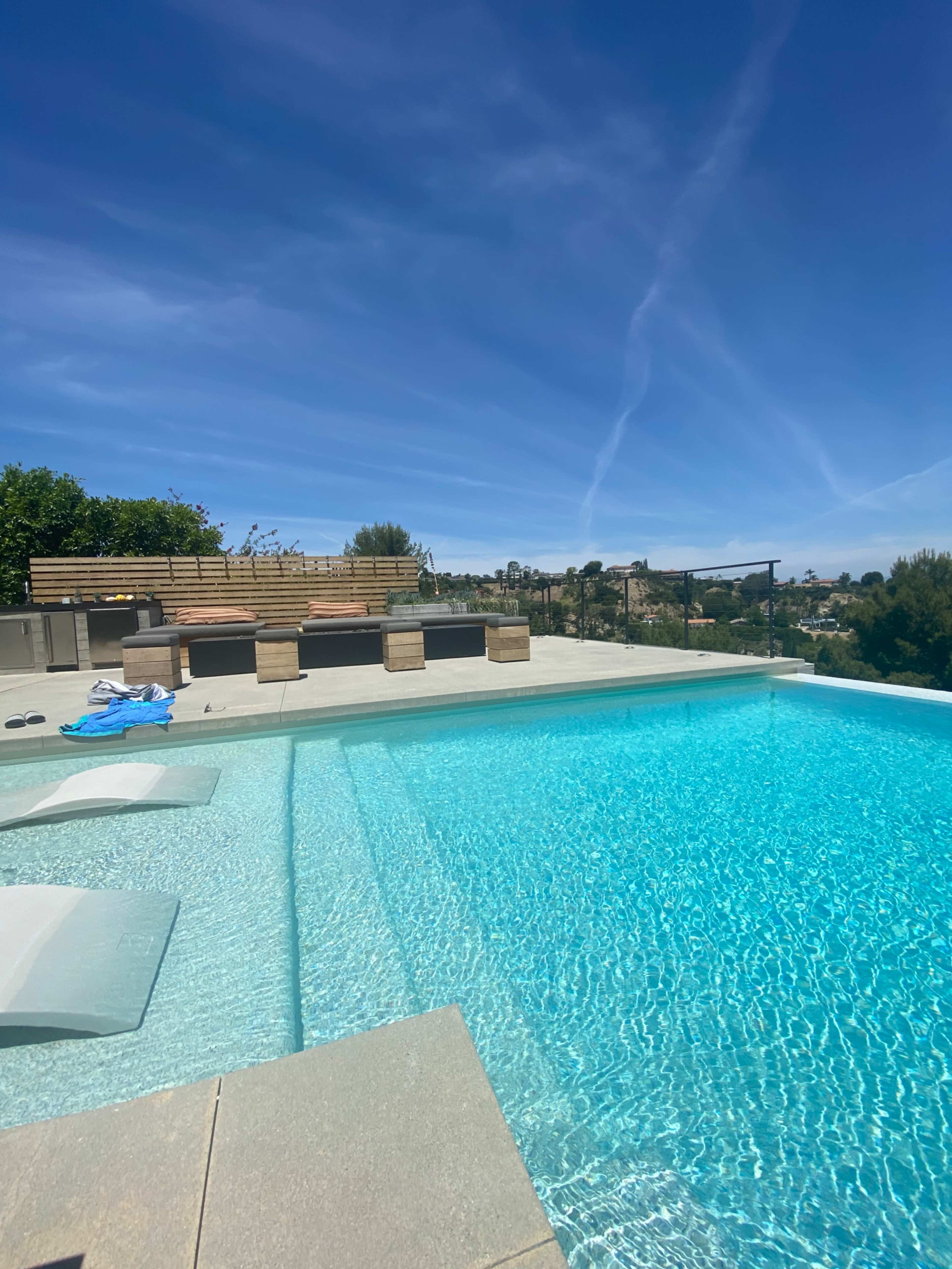 A clear swimming pool with a sunbathing area and lounge chairs against a backdrop of blue skies and distant hills.