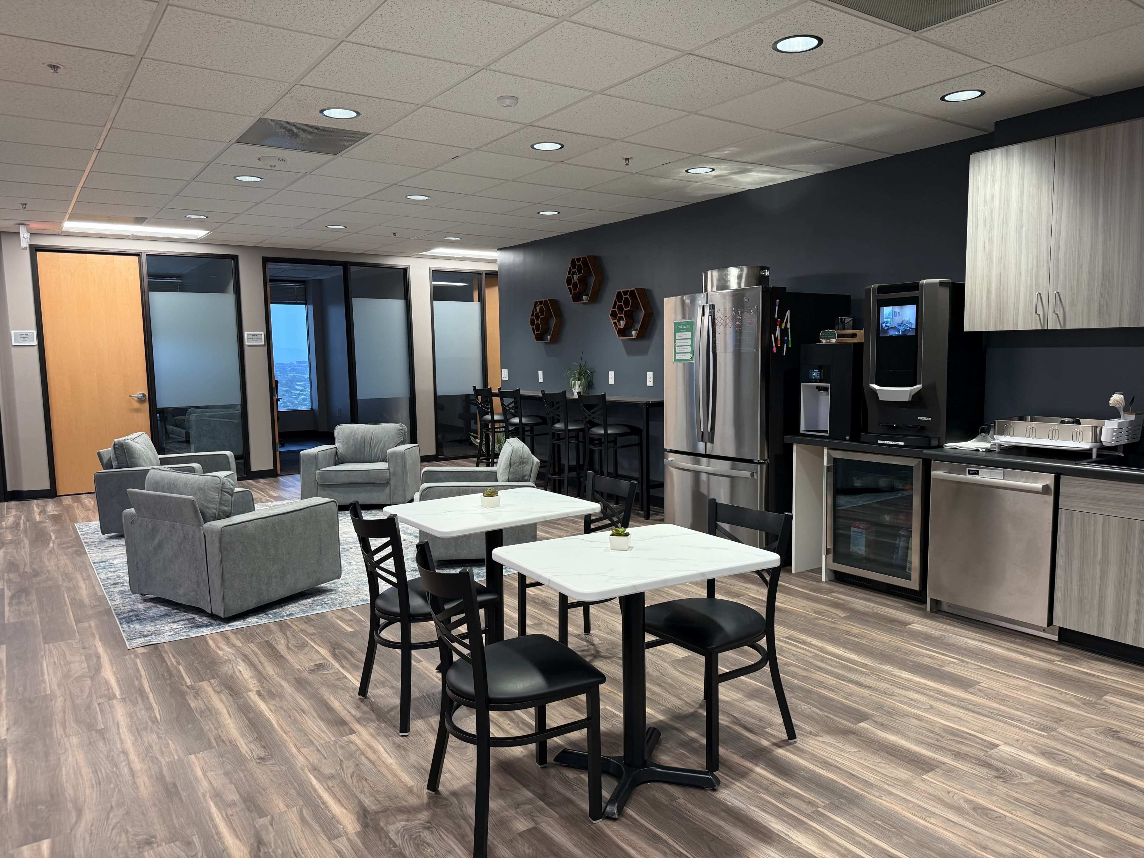 The image shows a modern break room with seating areas, tables, and kitchen appliances in an office environment.
