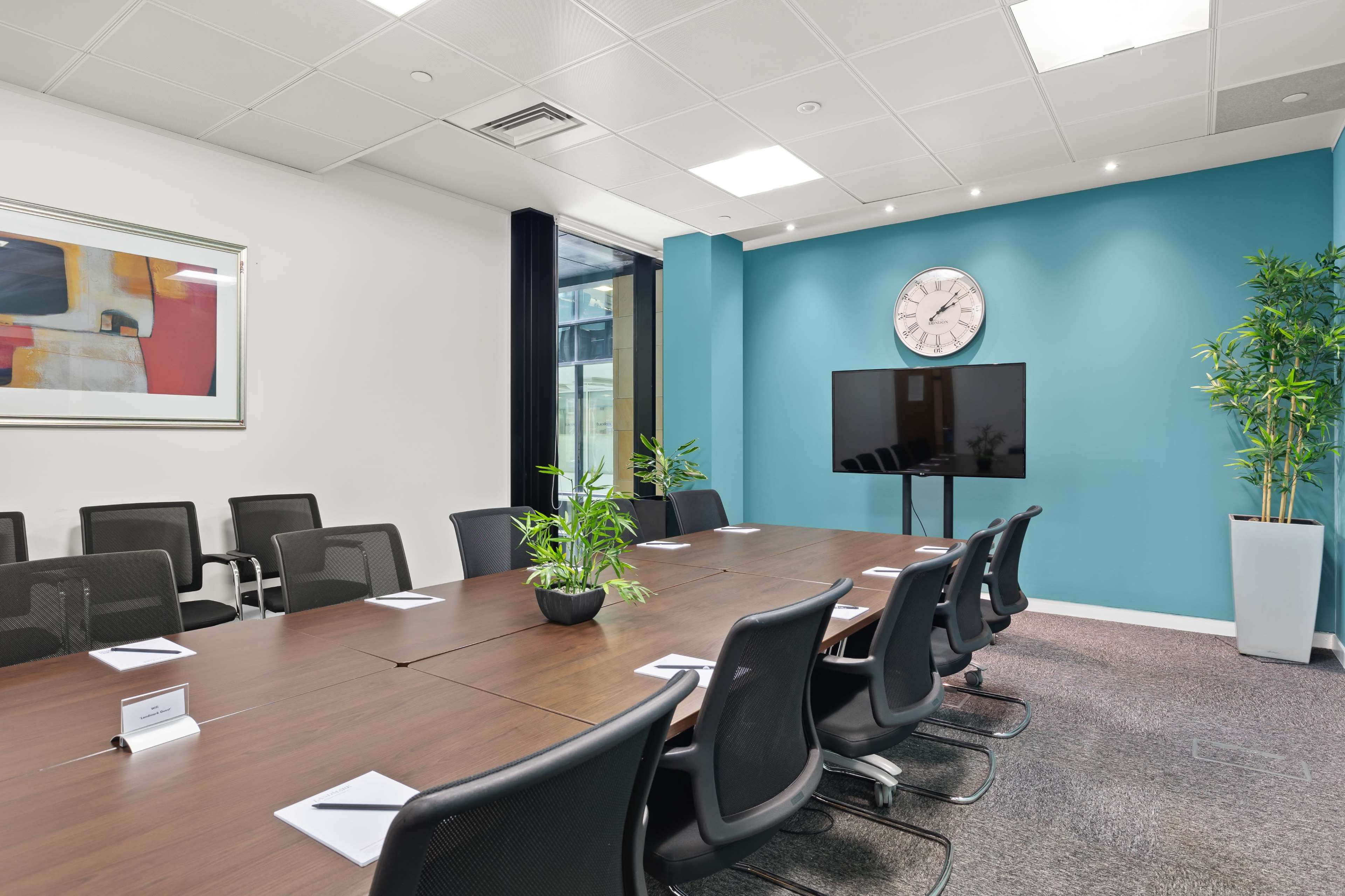 A conference room features a large wooden table surrounded by black chairs, a clock on the wall, a television monitor, and potted plants.