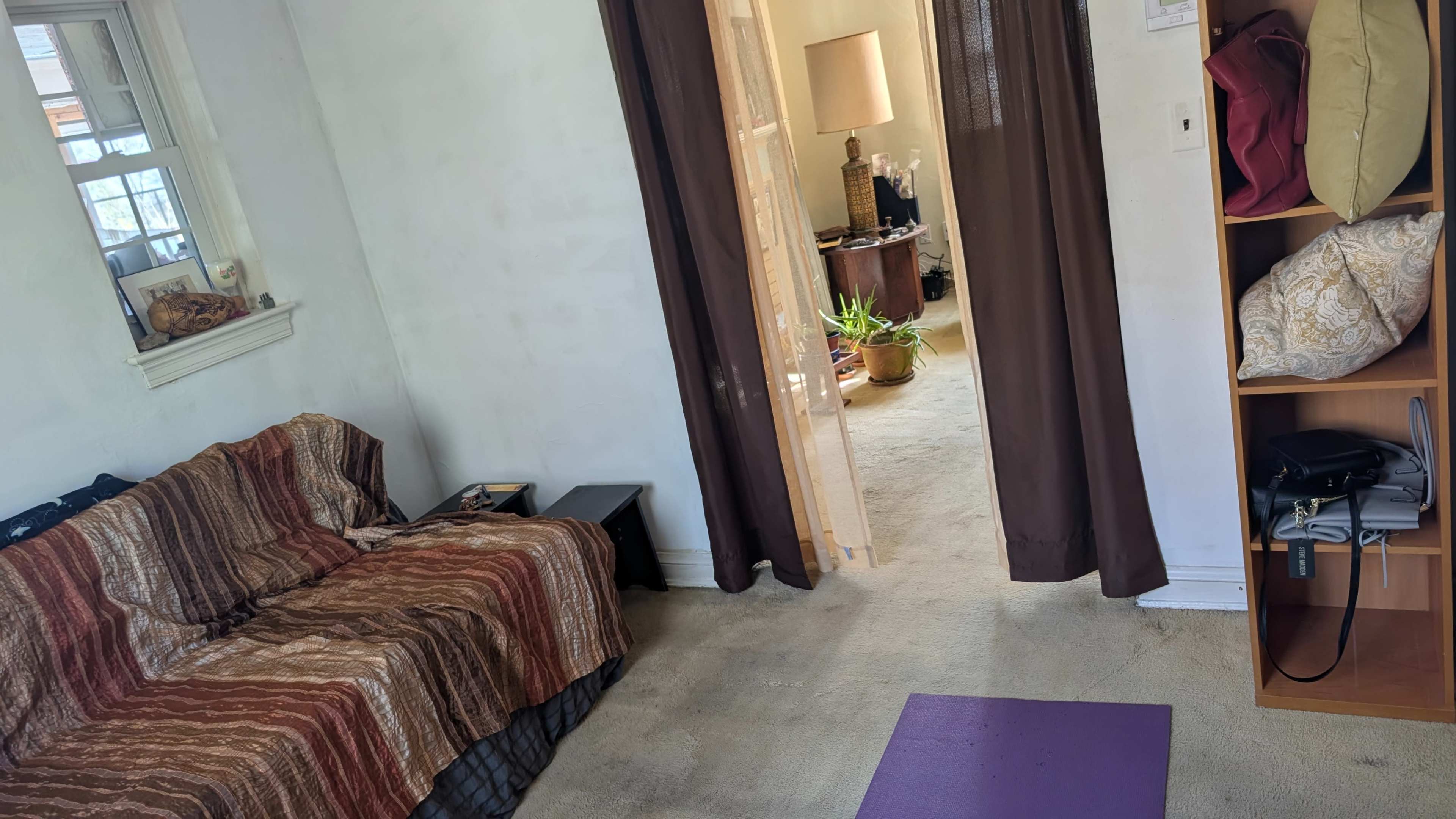 The image shows a modest living space with a patterned sofa, an open doorway leading to another room, and a yoga mat on the carpeted floor.