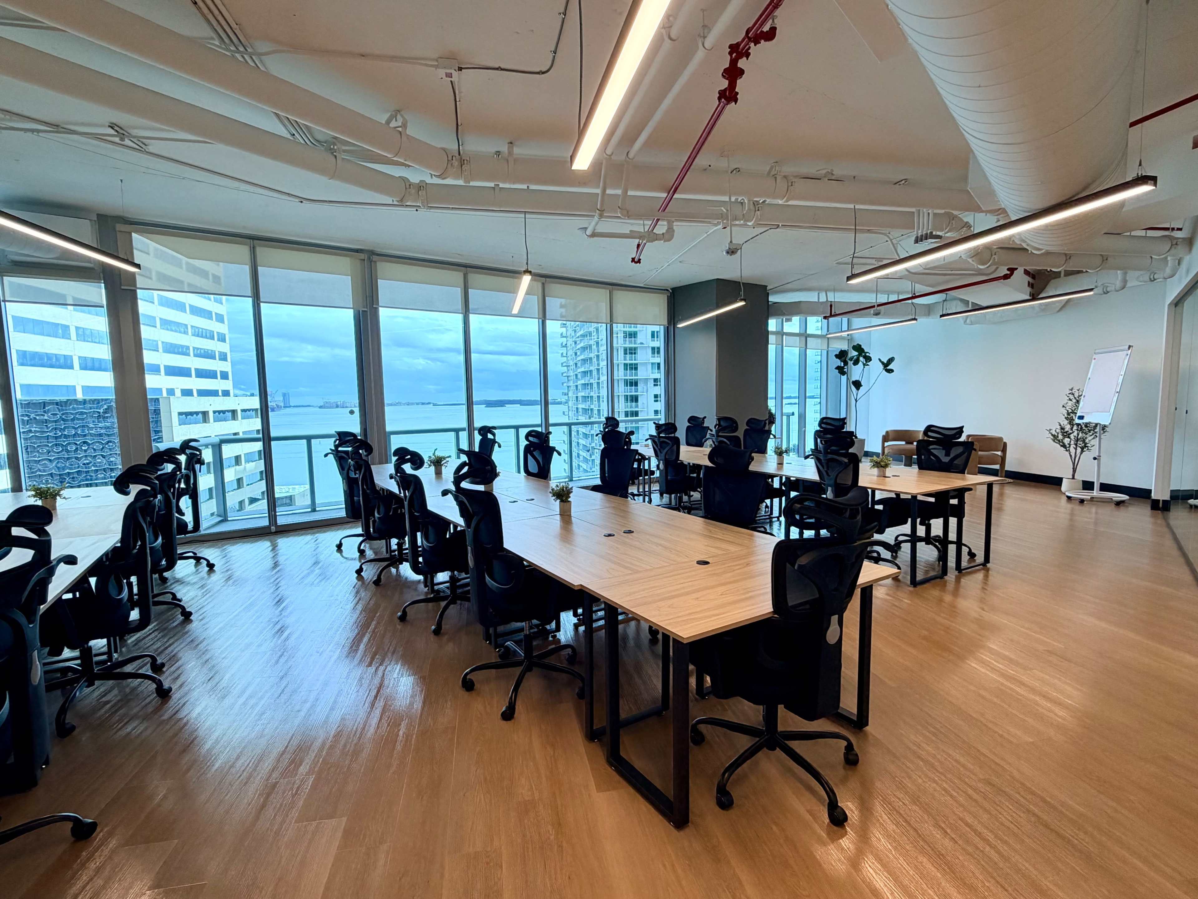 The image shows an empty office space with several tables and chairs arranged in a modern layout, large windows offering a view of a body of water and cityscape in the background.