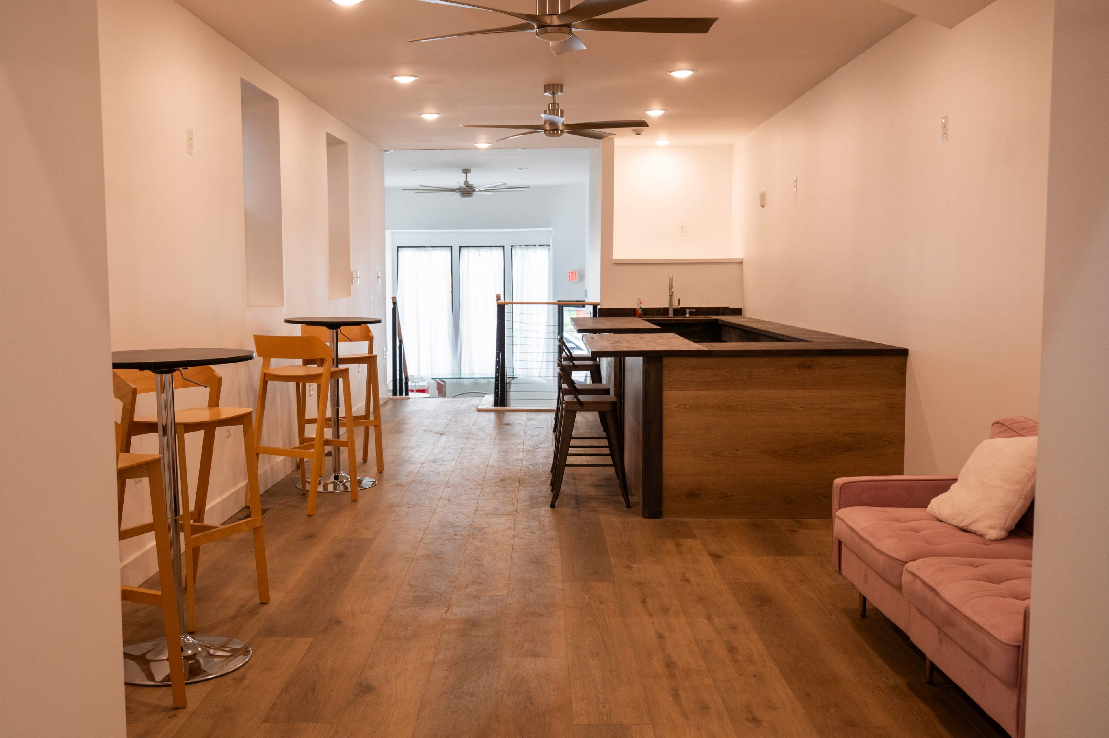 The image shows a spacious interior room featuring a bar area, several tall chairs, and a pink sofa, with natural light coming from windows at the far end.